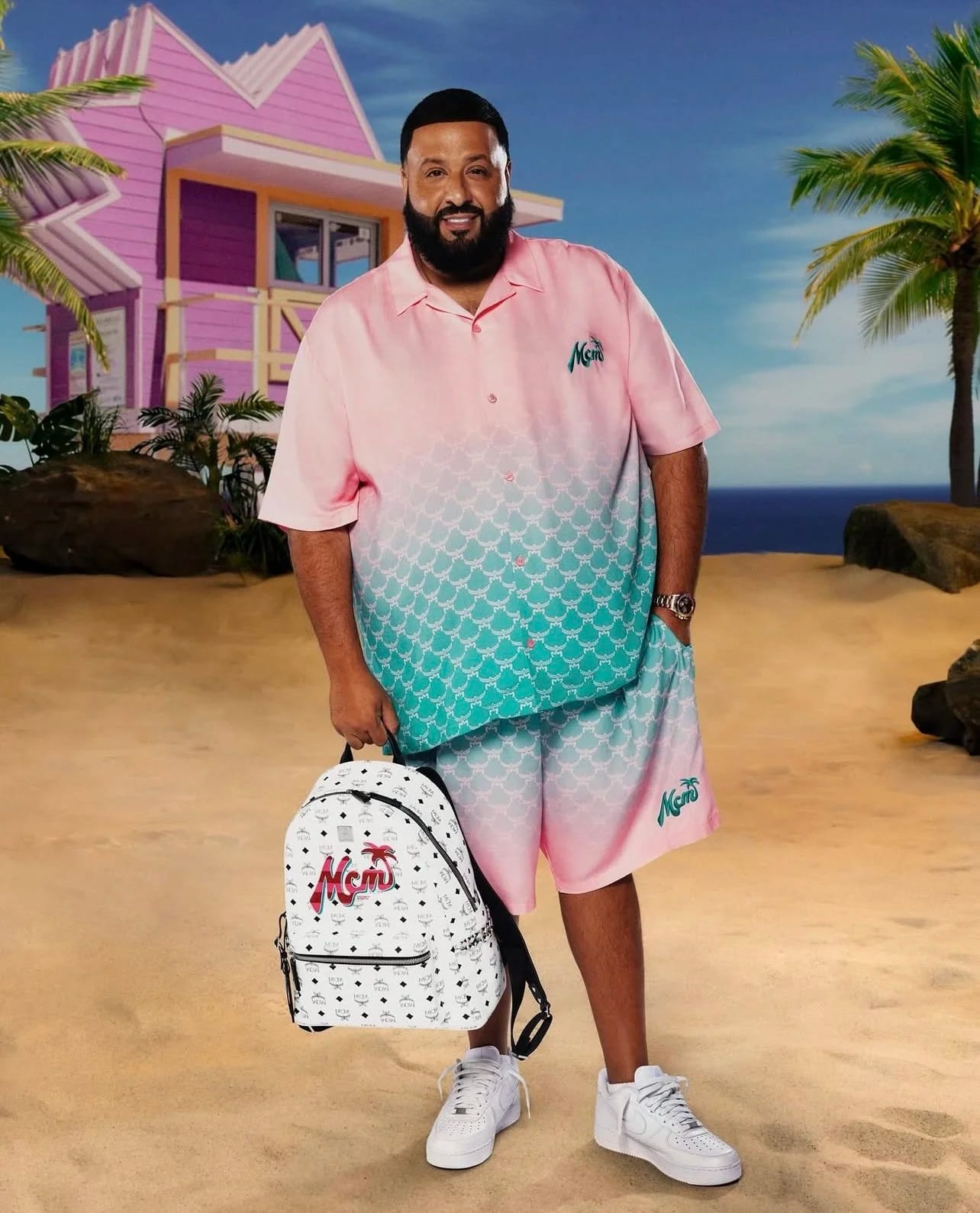 A bearded man in a pink and turquoise gradient shirt and shorts holding a white backpack on a beach set with a pink house and palms.