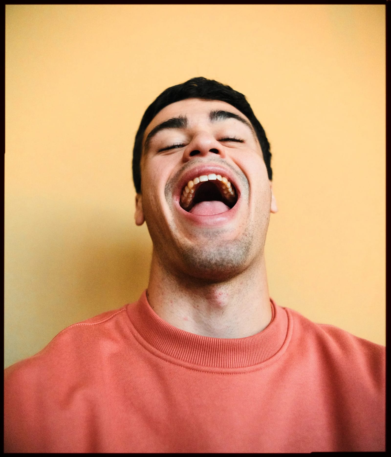 A young man in a coral sweatshirt laughing open-mouthed with his head tipped back against an orange-yellow wall.