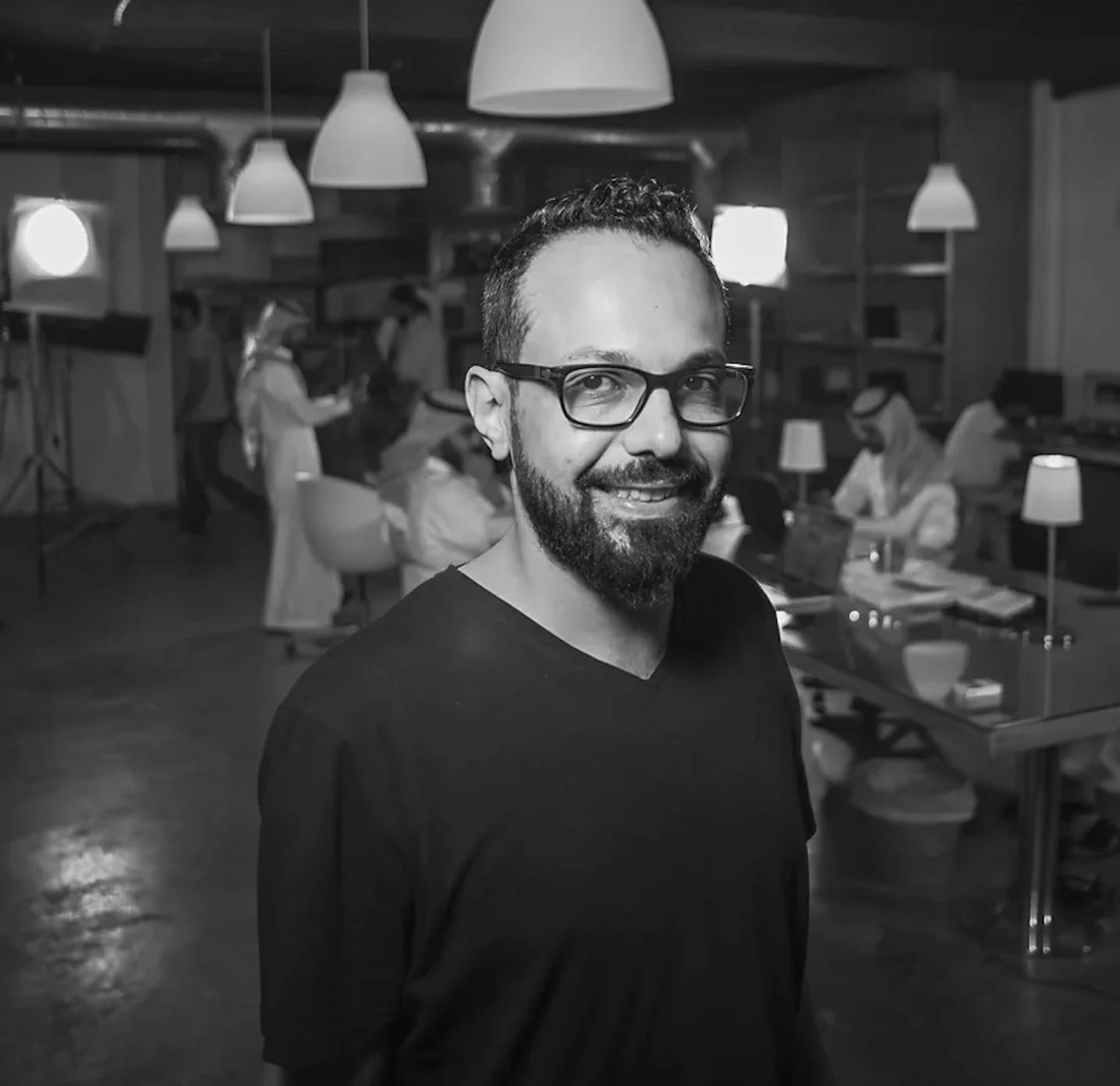 Black and white photograph of Mohammed Sendi in glasses and a black V-neck standing in a busy office with hanging lamps.