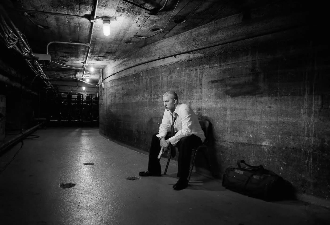 Black and white photograph of a man in a white shirt sitting on a chair in a long concrete tunnel, a bag on the floor beside him.