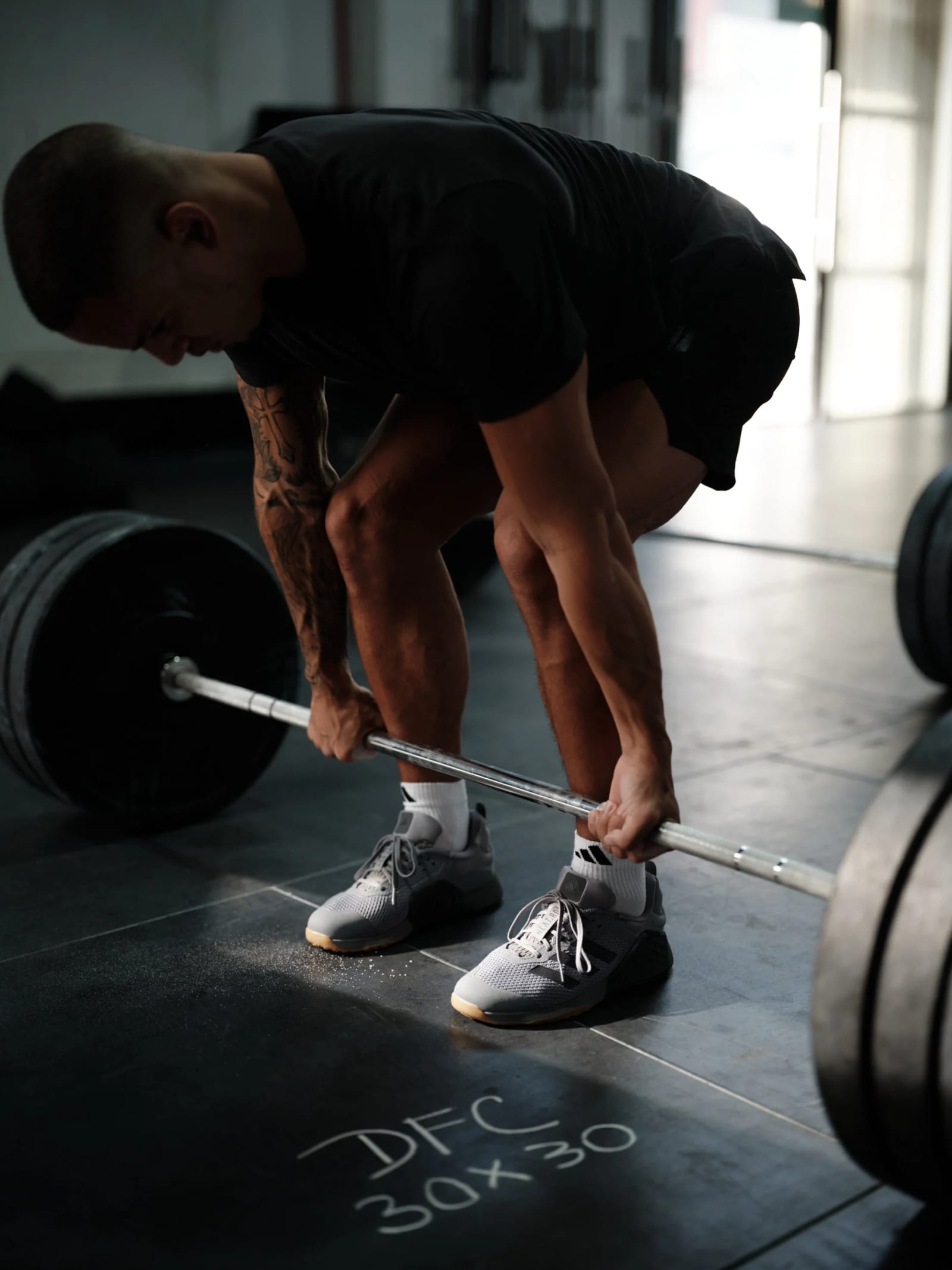A man in black shorts and a black T-shirt bending to grip a loaded barbell, a chalk note reading DFC 30x30 on the floor beside his shoes.