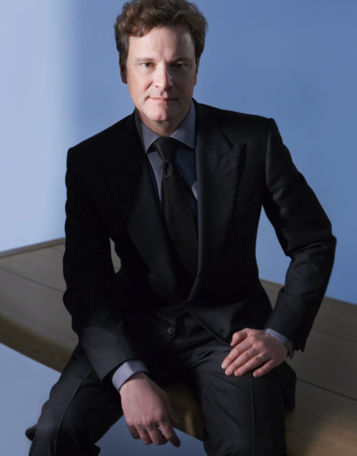 A man in a black suit sitting on the edge of a wooden table against a blue background.