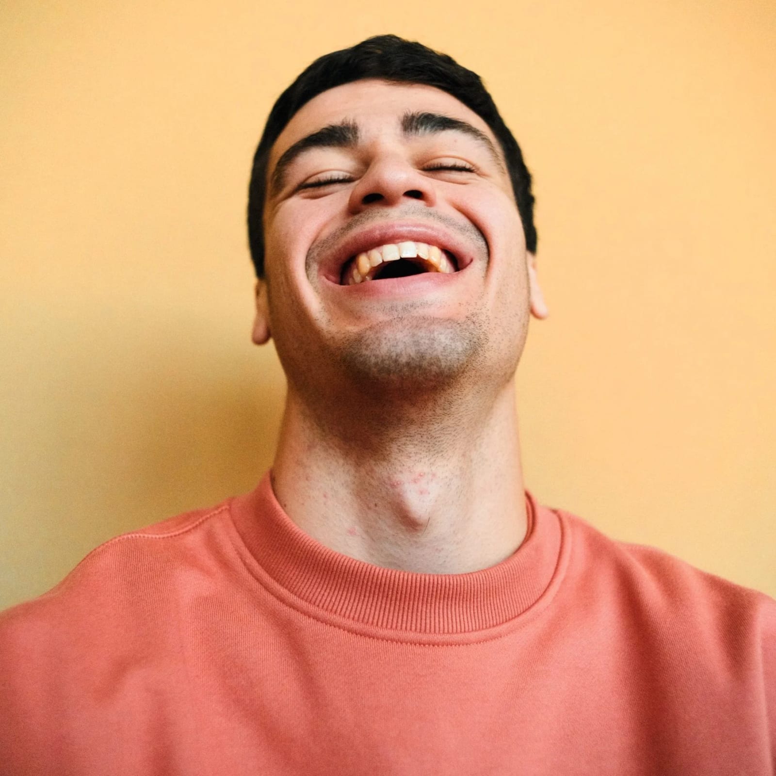 A young man in a coral sweatshirt laughing with his head tipped back against an orange-yellow wall.