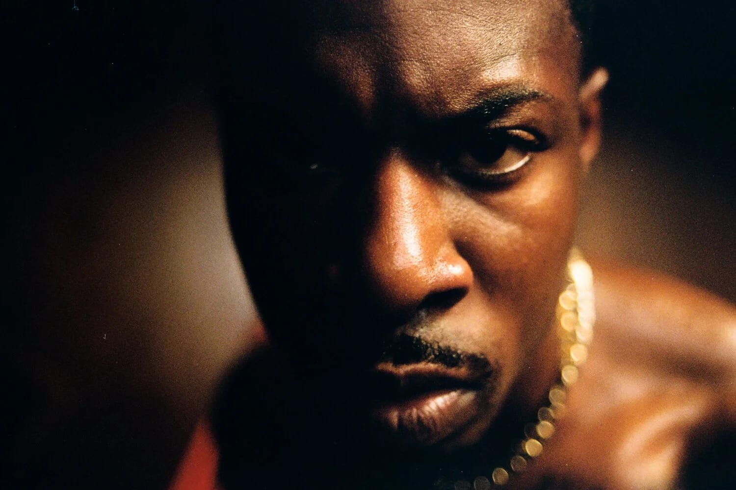 Close-up of a man's face in warm light with a gold chain at his neck, his eyes fixed on the camera.