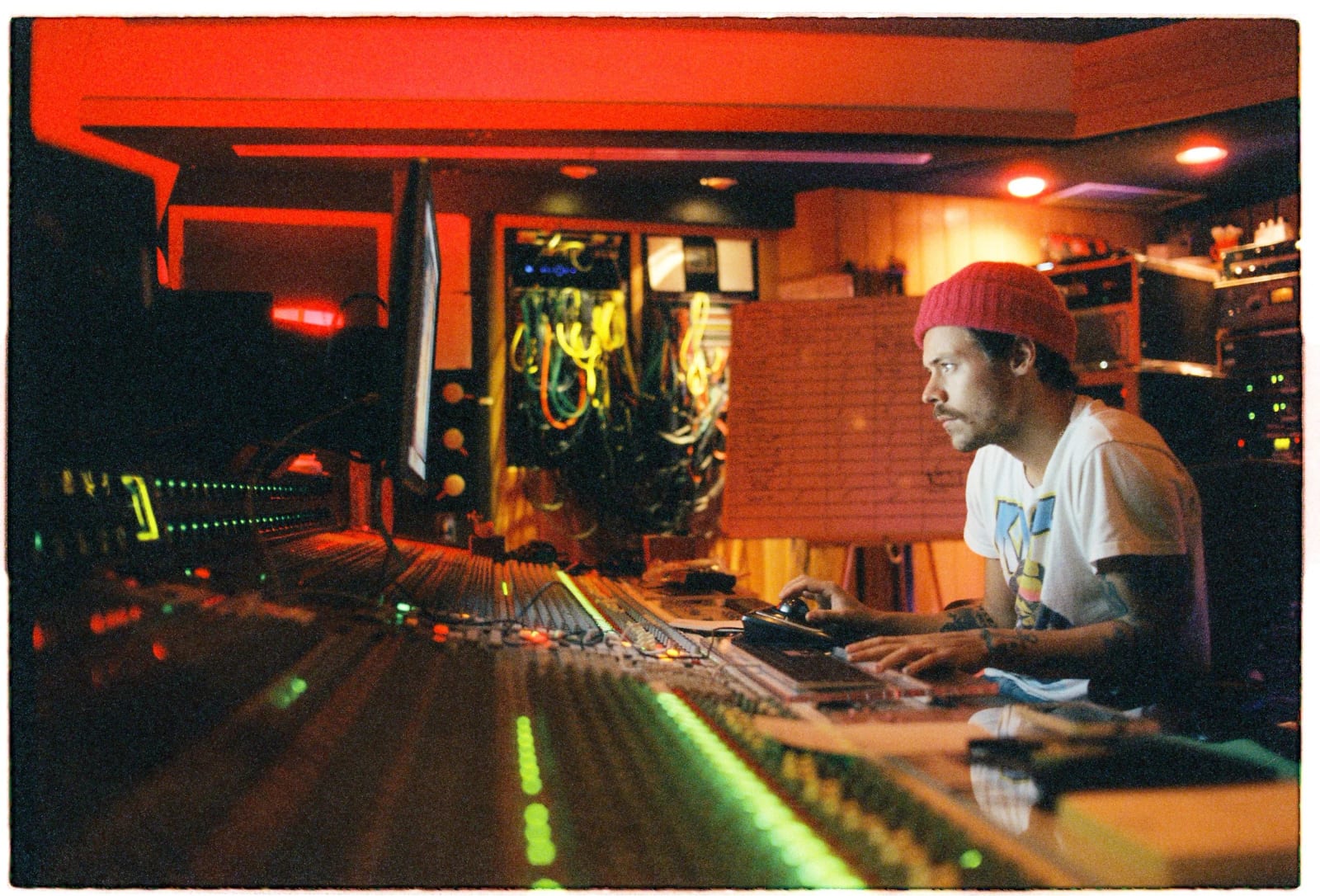 Harry Styles in a red beanie and white T-shirt at a mixing desk in a recording studio lit red, sheet music pinned above the console.