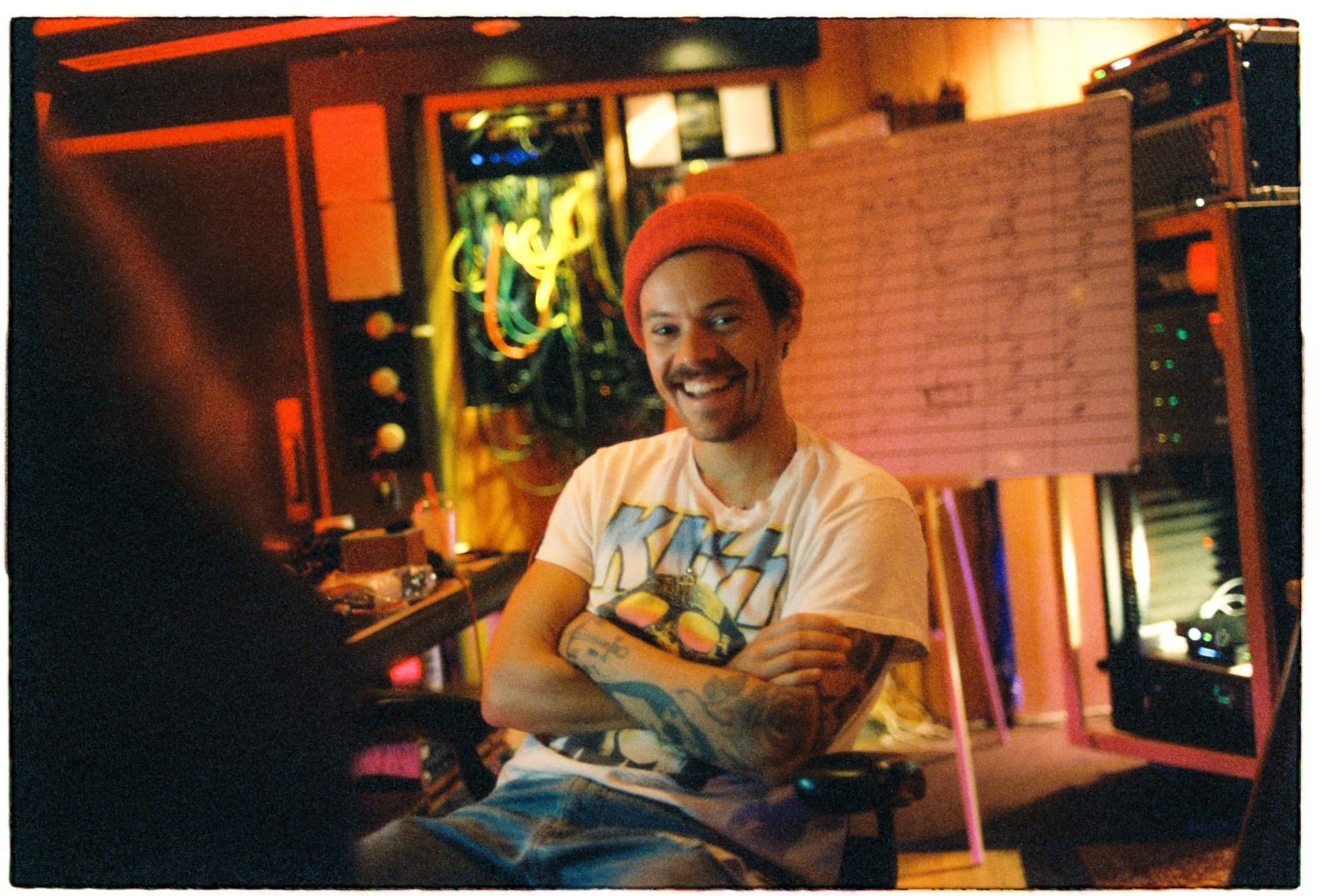 Harry Styles in a red beanie grinning with his arms folded in a red-lit recording studio.