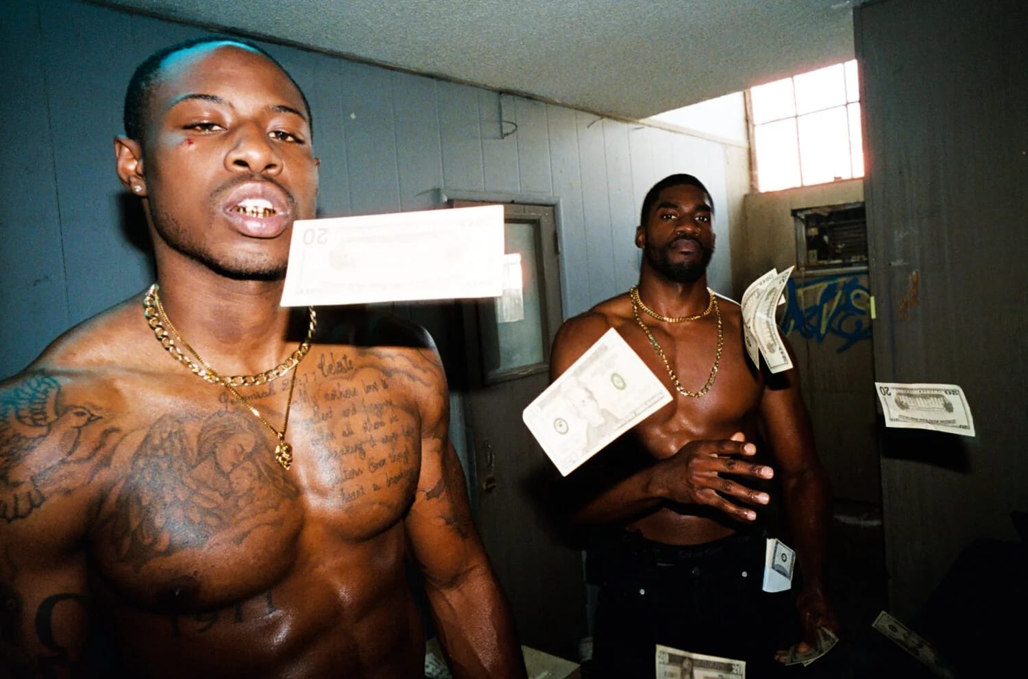 Two shirtless men in a bare room, one holding a banknote to the camera and the other with money in his hand.