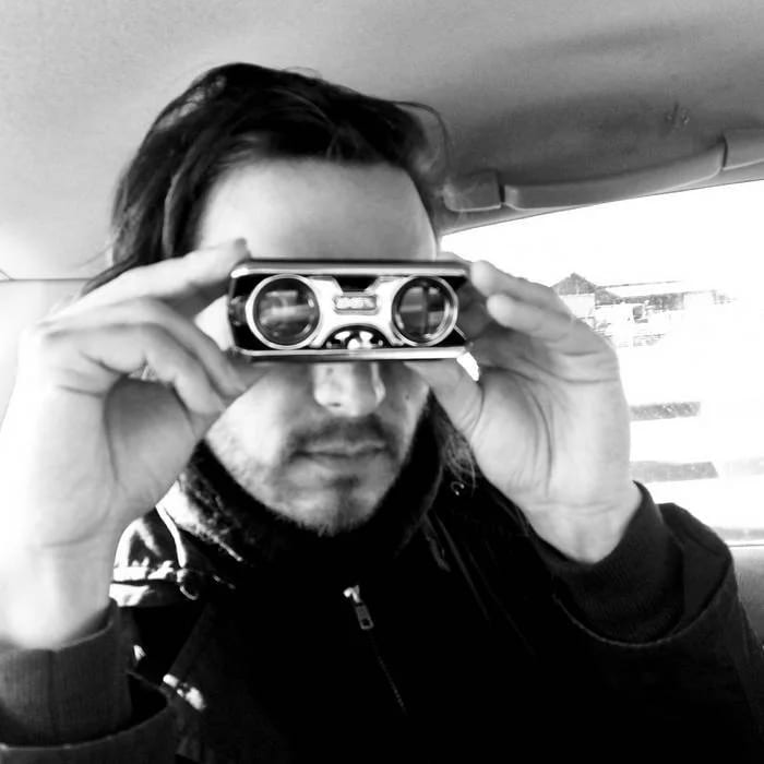Black and white photograph of Glassface holding small binoculars to his eyes inside a car.