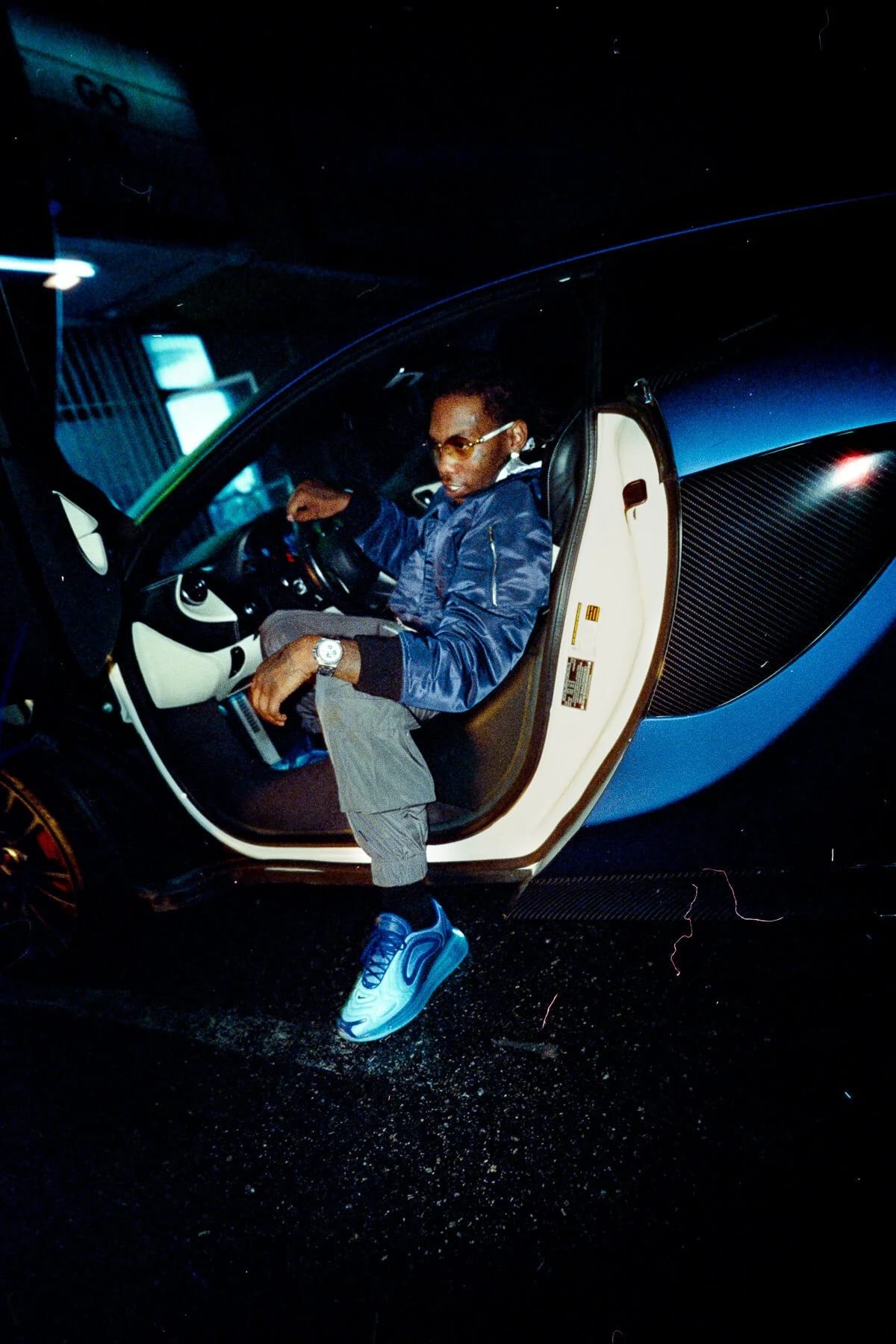 A man in sunglasses and a blue jacket sitting in the open door of a sports car in a dim garage, blue trainers on the ground.