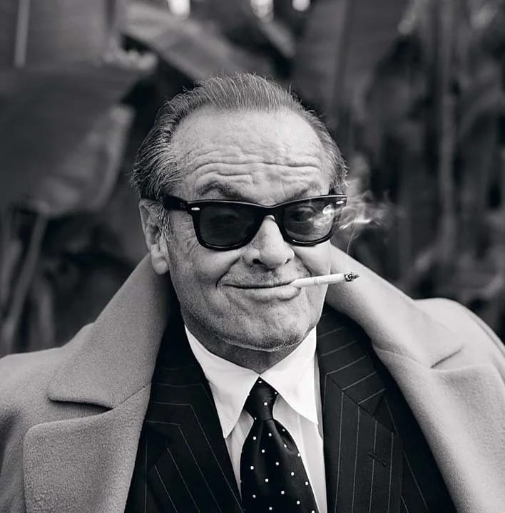 Black and white portrait of Jack Nicholson in sunglasses and a pinstripe suit with a cigarette, in front of palm leaves.