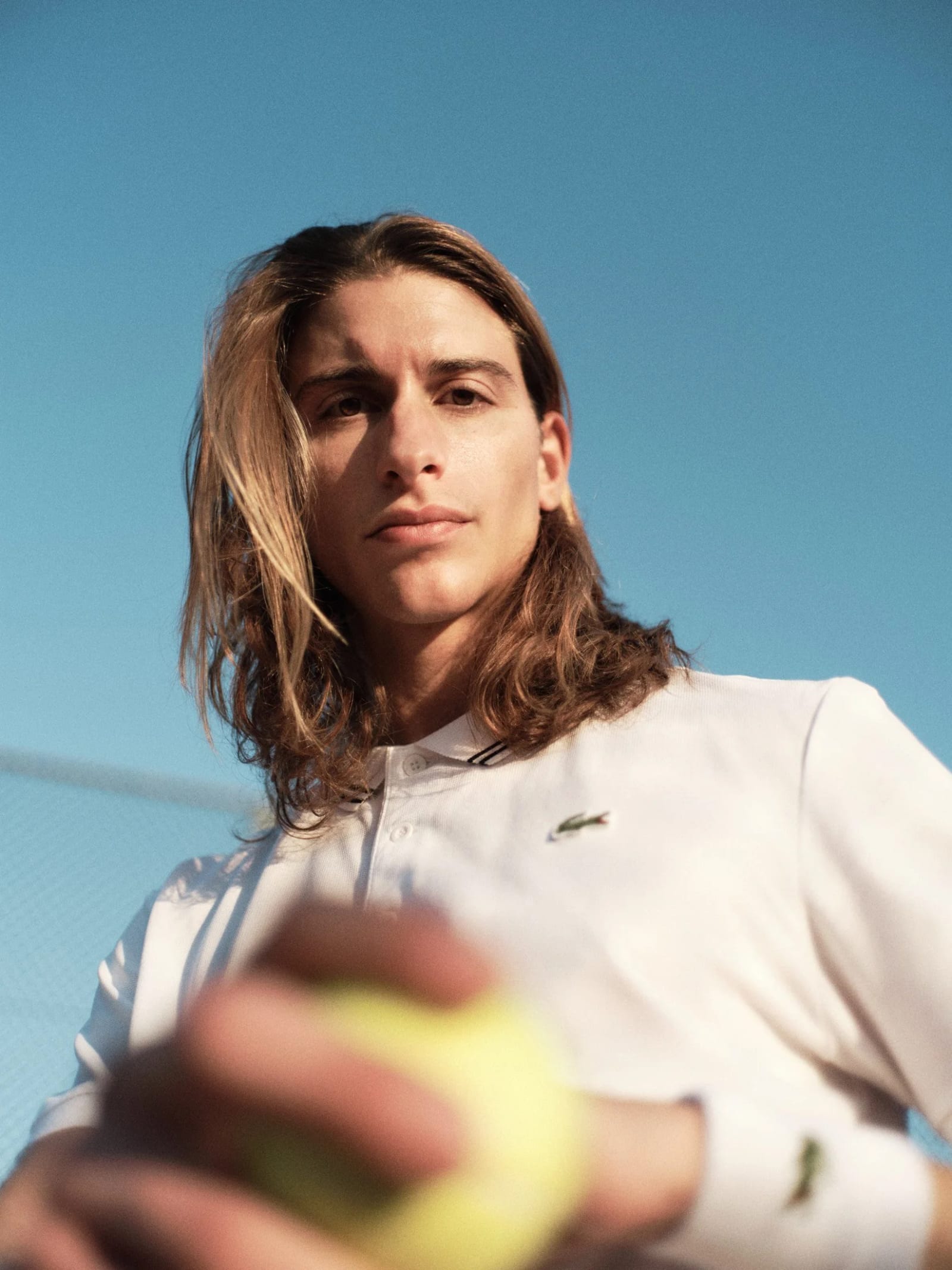 A long-haired man in a white Lacoste polo holding a tennis ball, photographed from below against a blue sky.