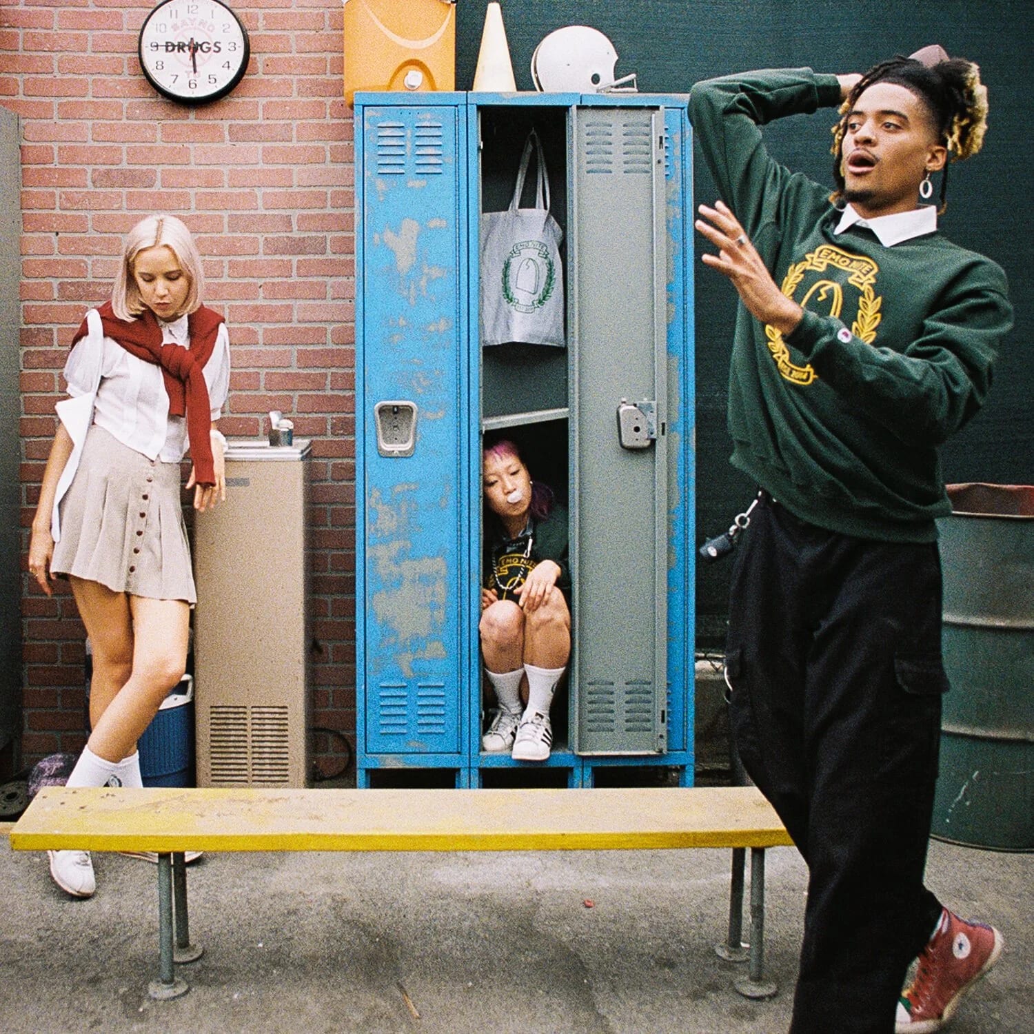 A man in a green sweatshirt dancing beside blue school lockers, one open with a woman sitting inside it, a woman in a skirt leaning on the wall.