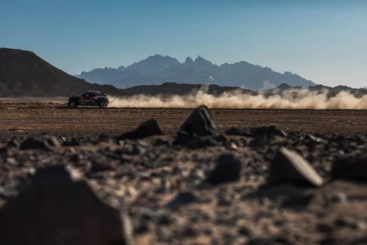 A rally car trailing dust across a stony desert plain, mountains rising behind.
