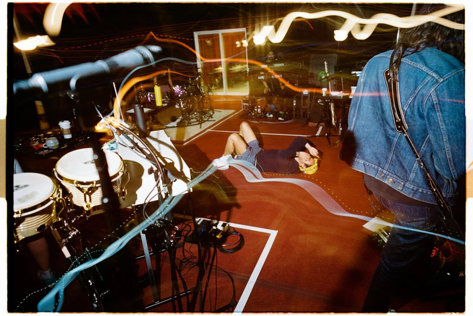 A guitarist in a denim jacket on a stage floor, streaks of light trailing across the frame from a long exposure.