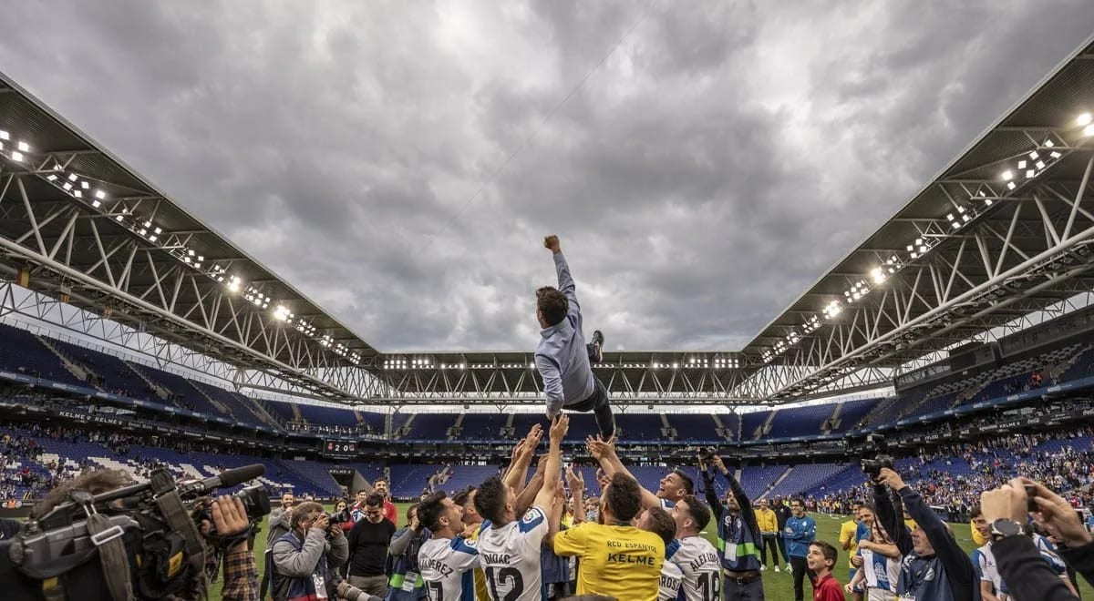 A footballer thrown into the air by a crowd of teammates on a stadium pitch under grey clouds.