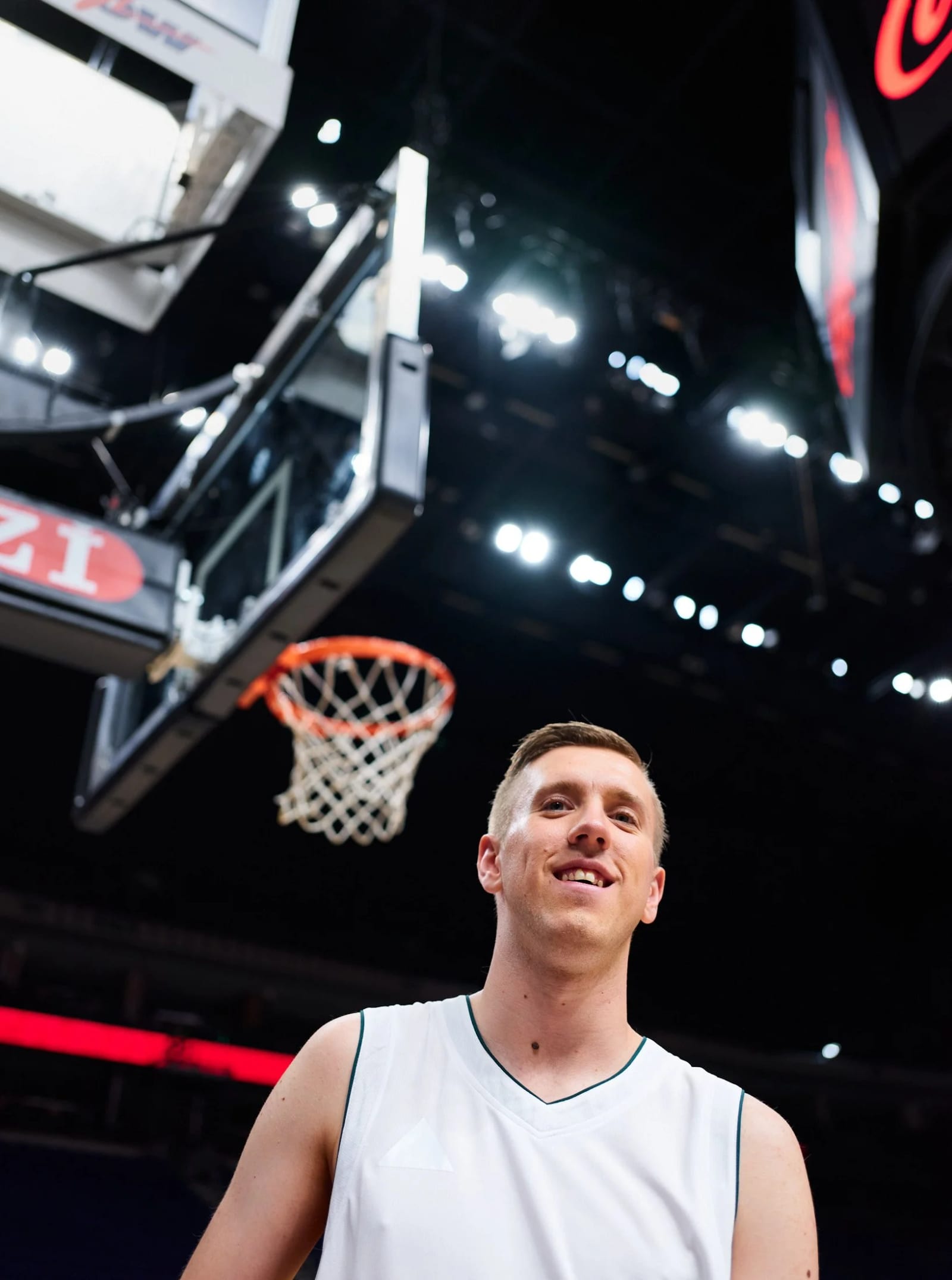 A smiling man in a white basketball jersey beneath a hoop, arena lights and a red Coca-Cola sign above him.