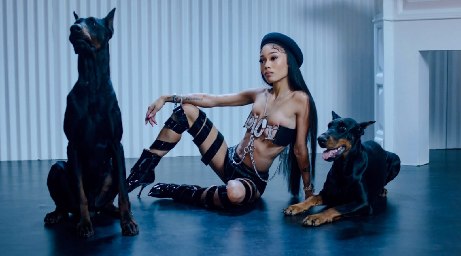 A woman in a black cap and strappy boots sitting on the floor between two Dobermans in a white room.