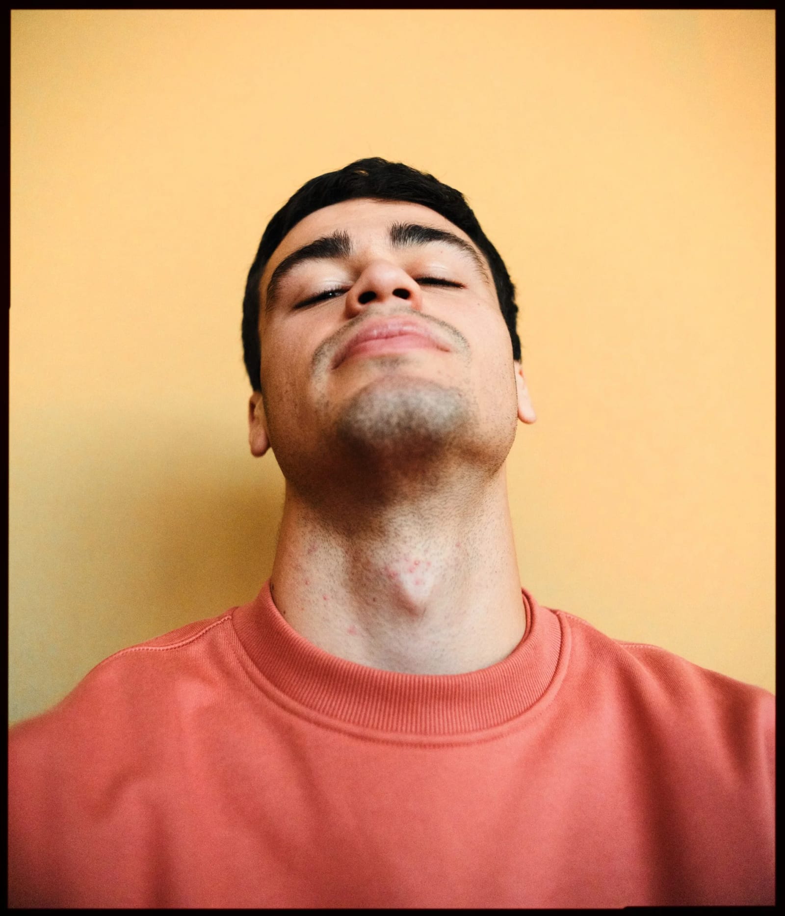 A young man in a coral sweatshirt with his eyes closed and chin raised against an orange-yellow wall.
