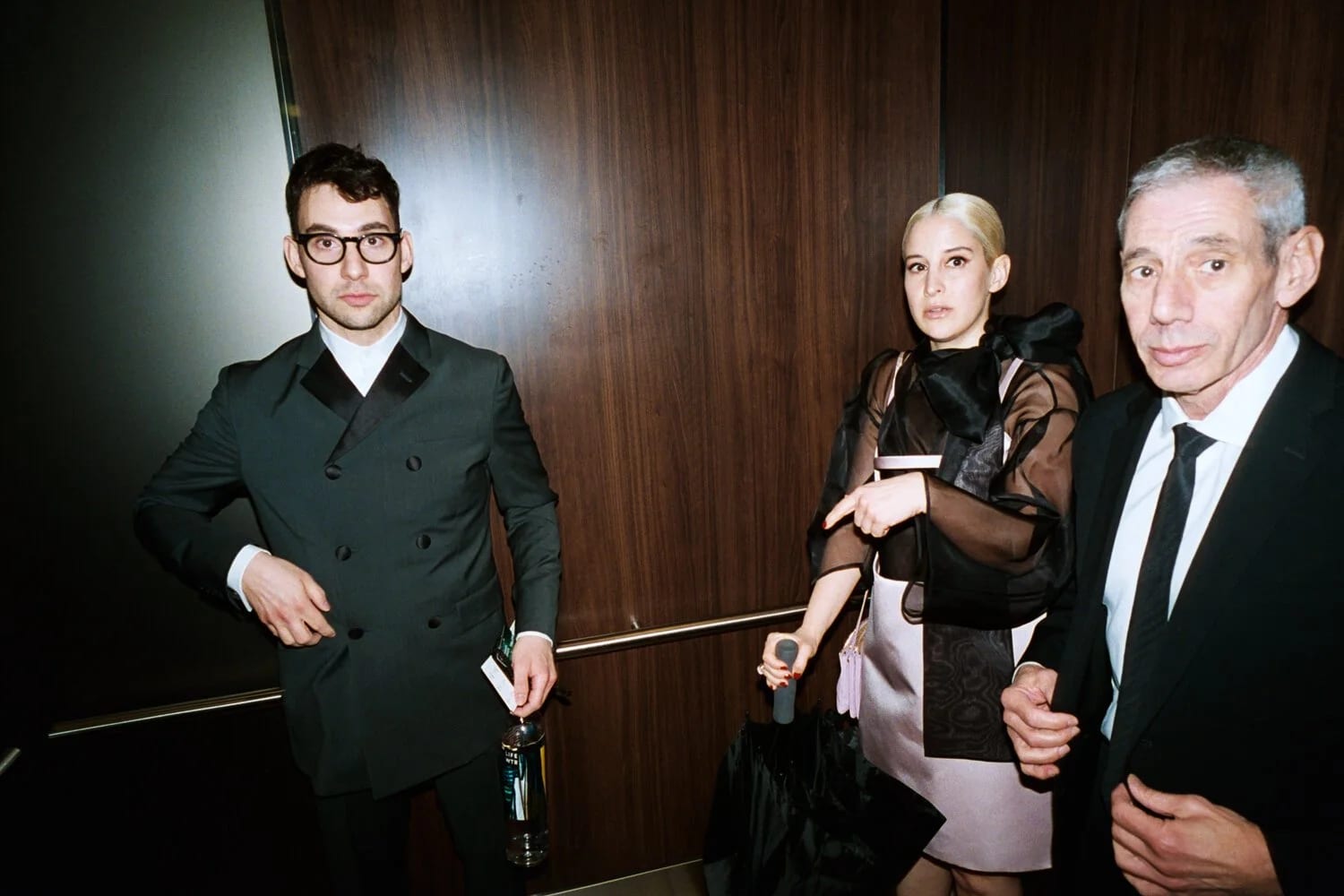 A man in glasses and a dark suit, a woman with platinum hair in a sheer top and an older man in a tie standing in a wood-panelled lift.