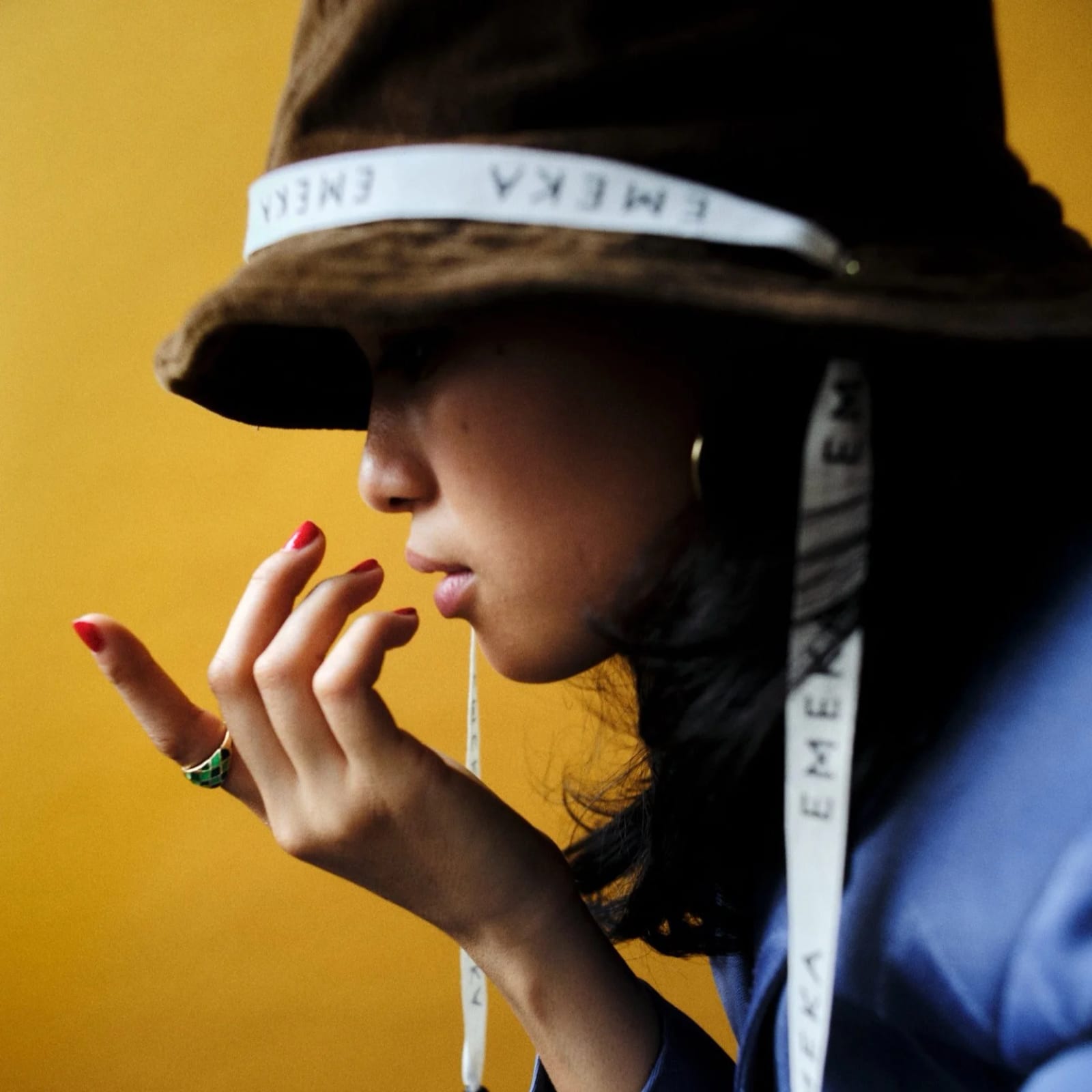 Profile of a woman in a brown bucket hat with a white strap lettered EMEKA, red-nailed fingers raised to her lips, against a yellow wall.