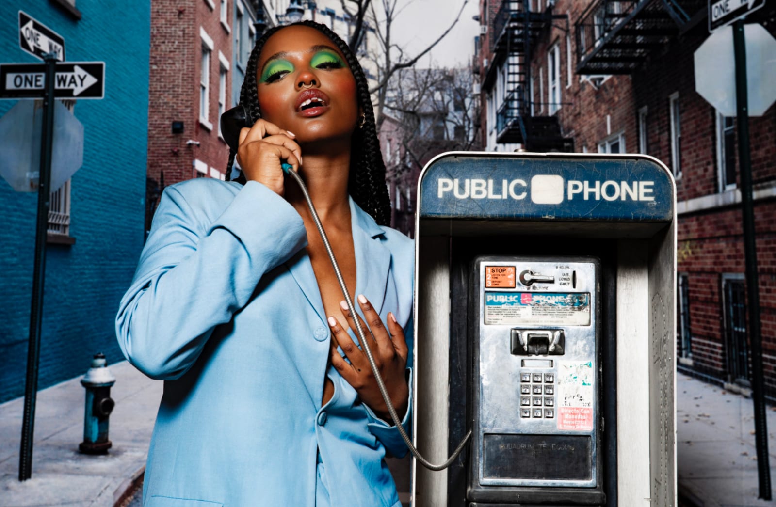 A woman with green eyeshadow in a pale blue suit talking on a public payphone on a city street.