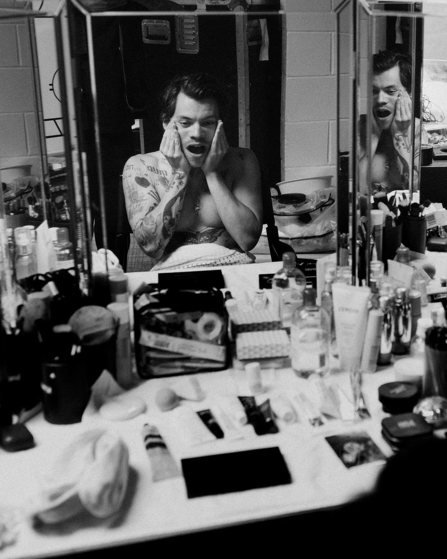 Black and white photograph of a shirtless Harry Styles holding his face at a dressing-room mirror crowded with bottles and make-up.