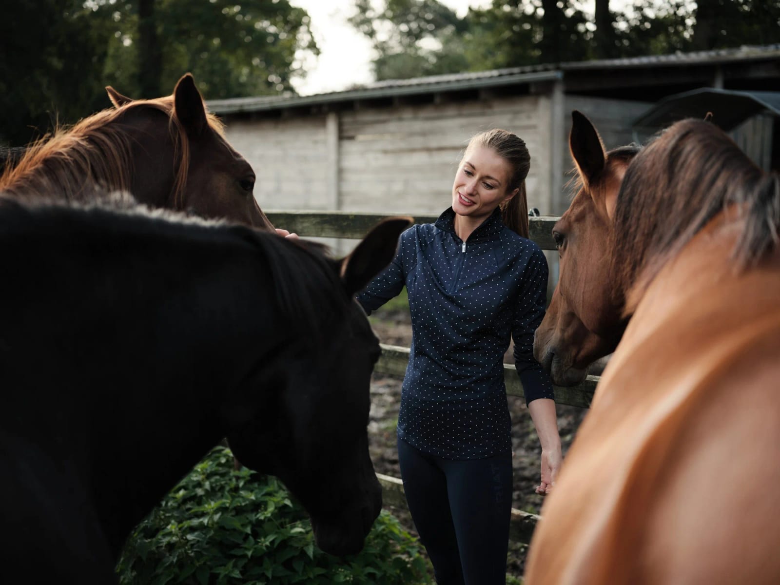 A smiling woman in a navy top between two chestnut horses in a stable yard.