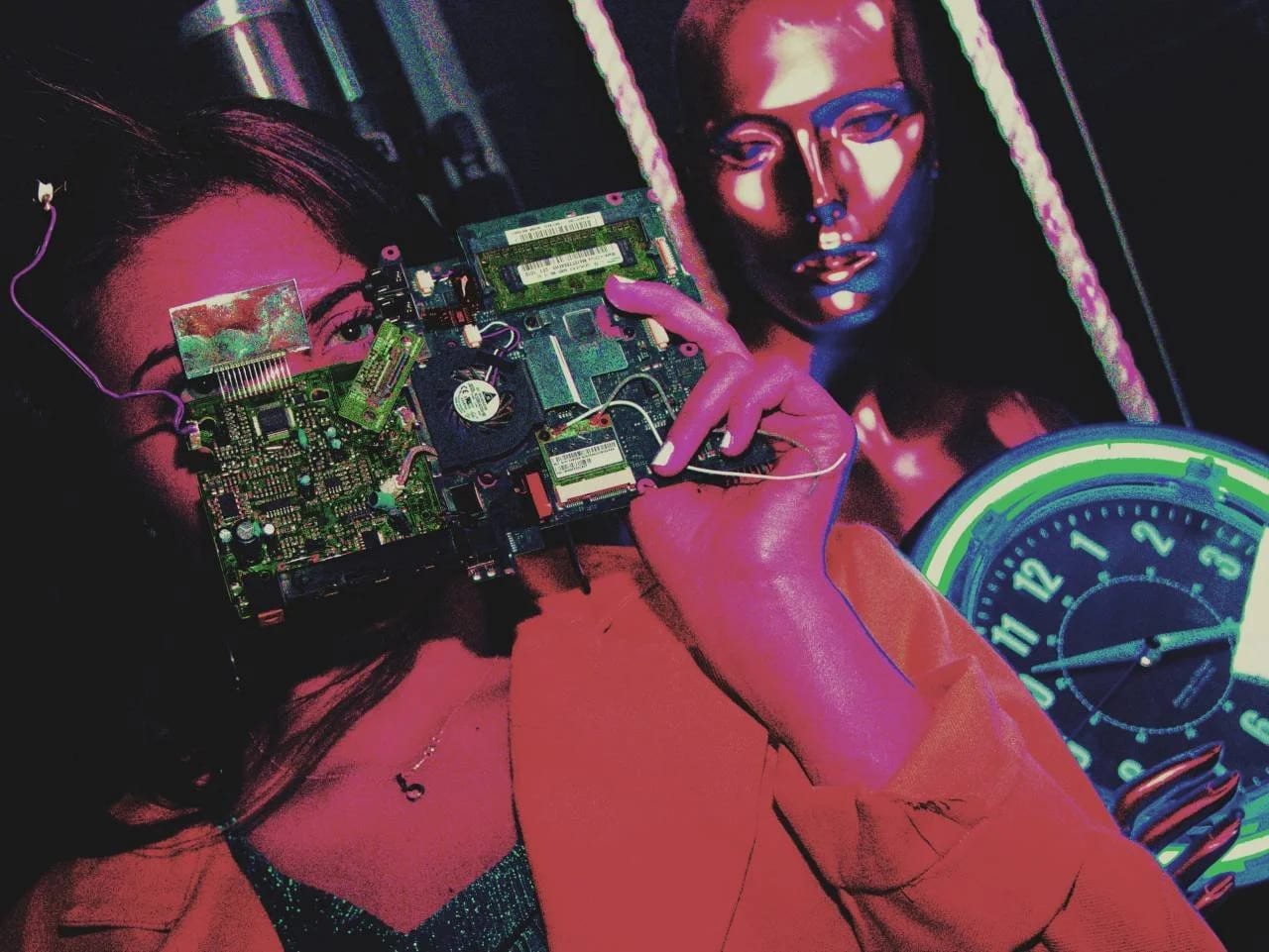 A woman holding a circuit board to her face beside a figure painted metallic bronze, a clock behind, in dim red light.