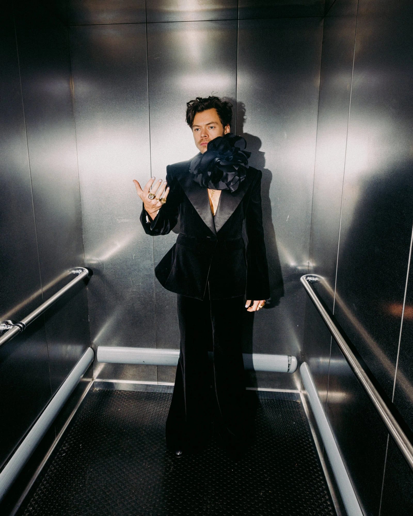 A man in a black velvet suit with a ruffled collar standing in a steel-walled lift, one hand held out.