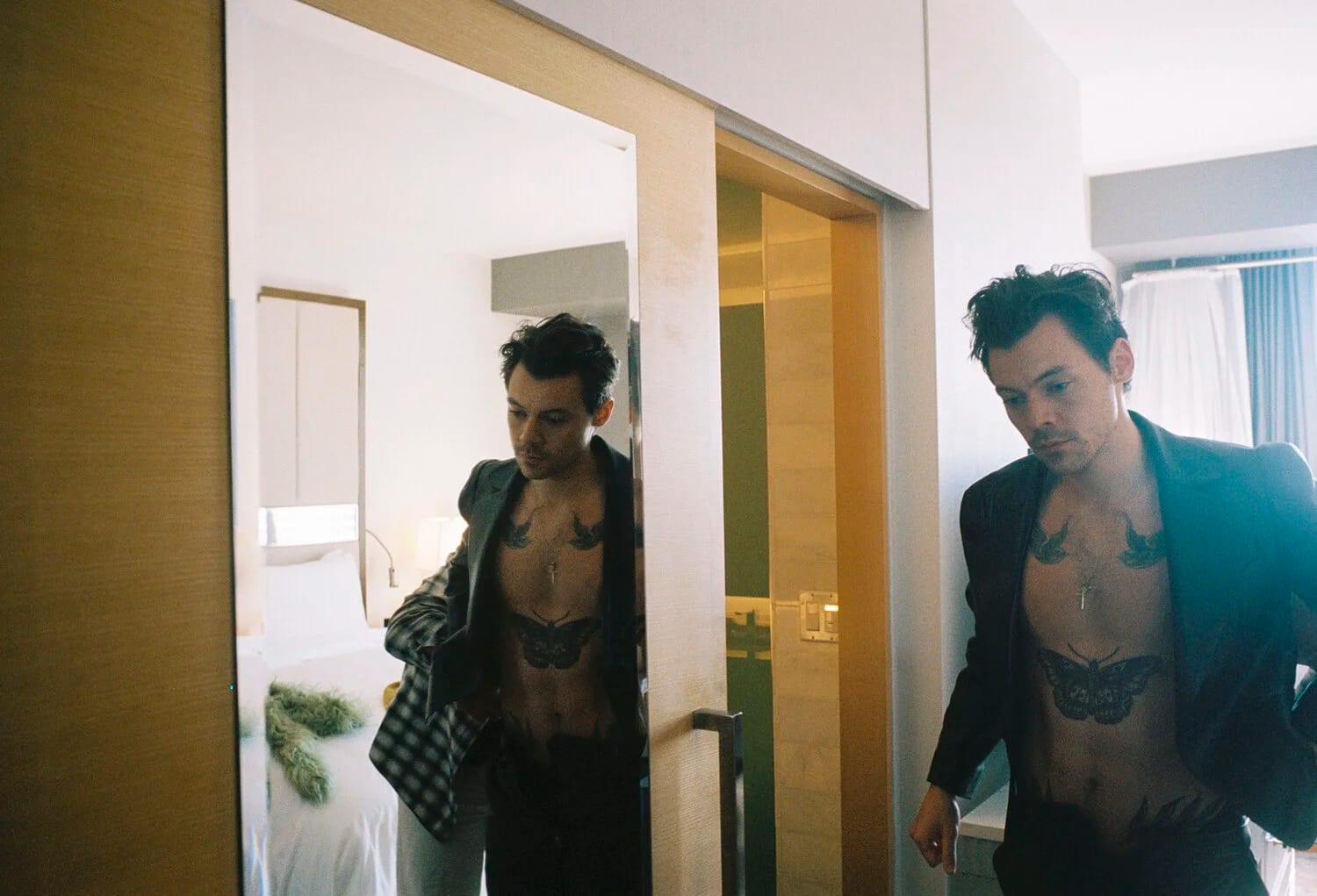 Harry Styles in an open black jacket seen twice, once in a mirror, standing in a hotel bathroom doorway.