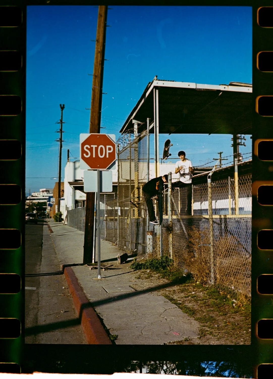A film frame of a stop sign and chain-link fence on a sunlit street corner, two people climbing the fence behind.