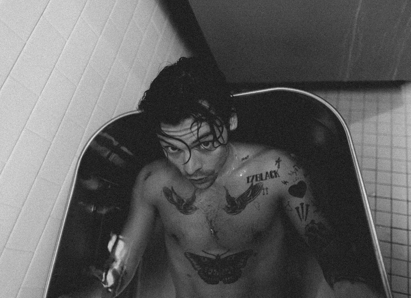 Black and white photograph of a tattooed Harry Styles in a bath, looking up at the camera with wet hair.
