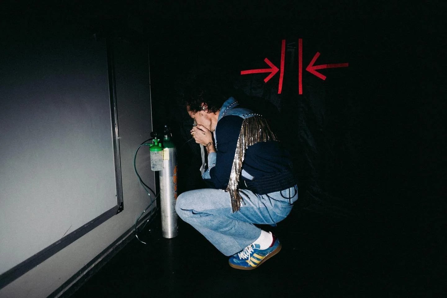 Harry Styles crouching against a black wall in a dark jacket and jeans, a hand over his face, red arrows painted above him.