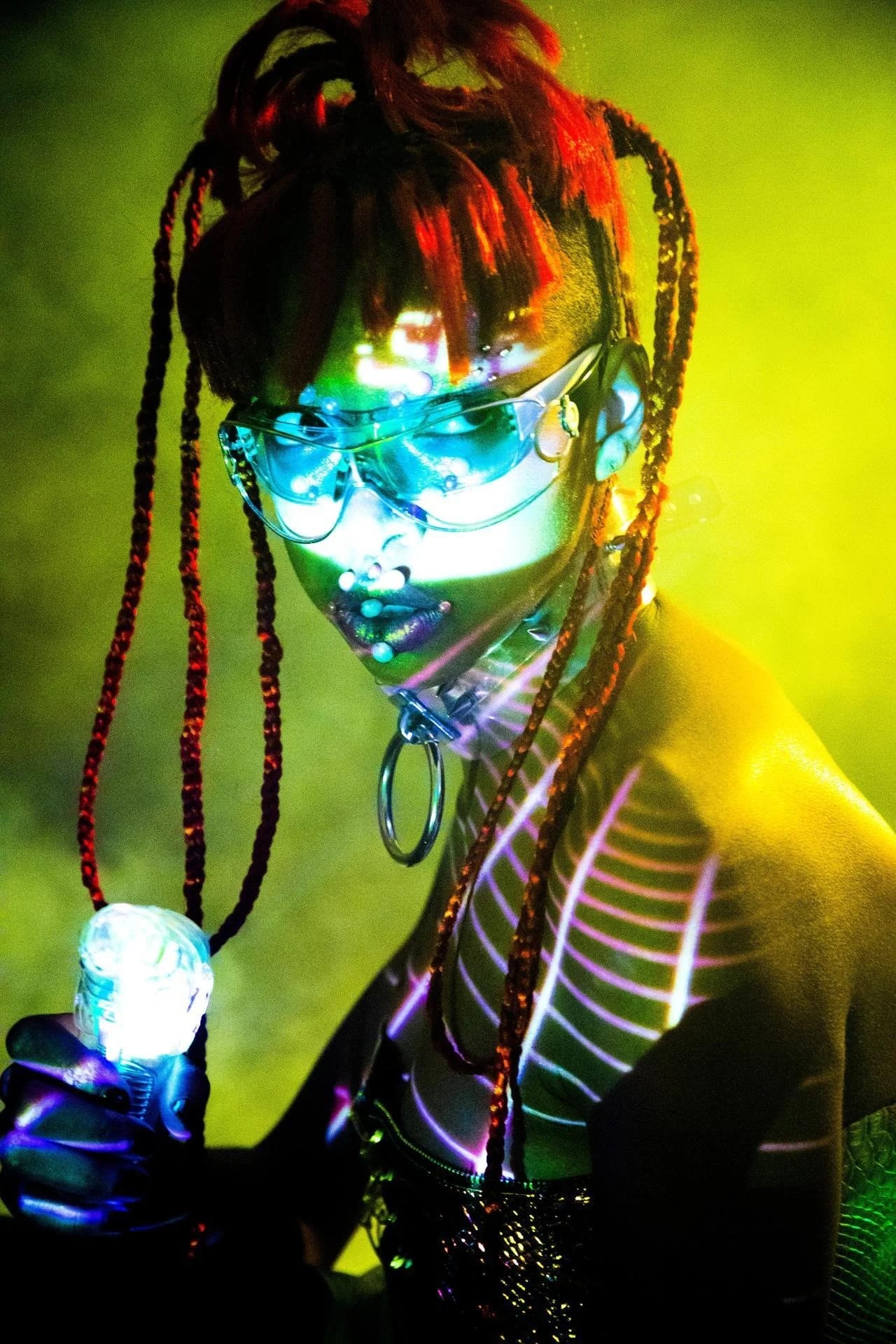 A person with long braids and mirrored glasses holding a glowing orb, lit green and red.