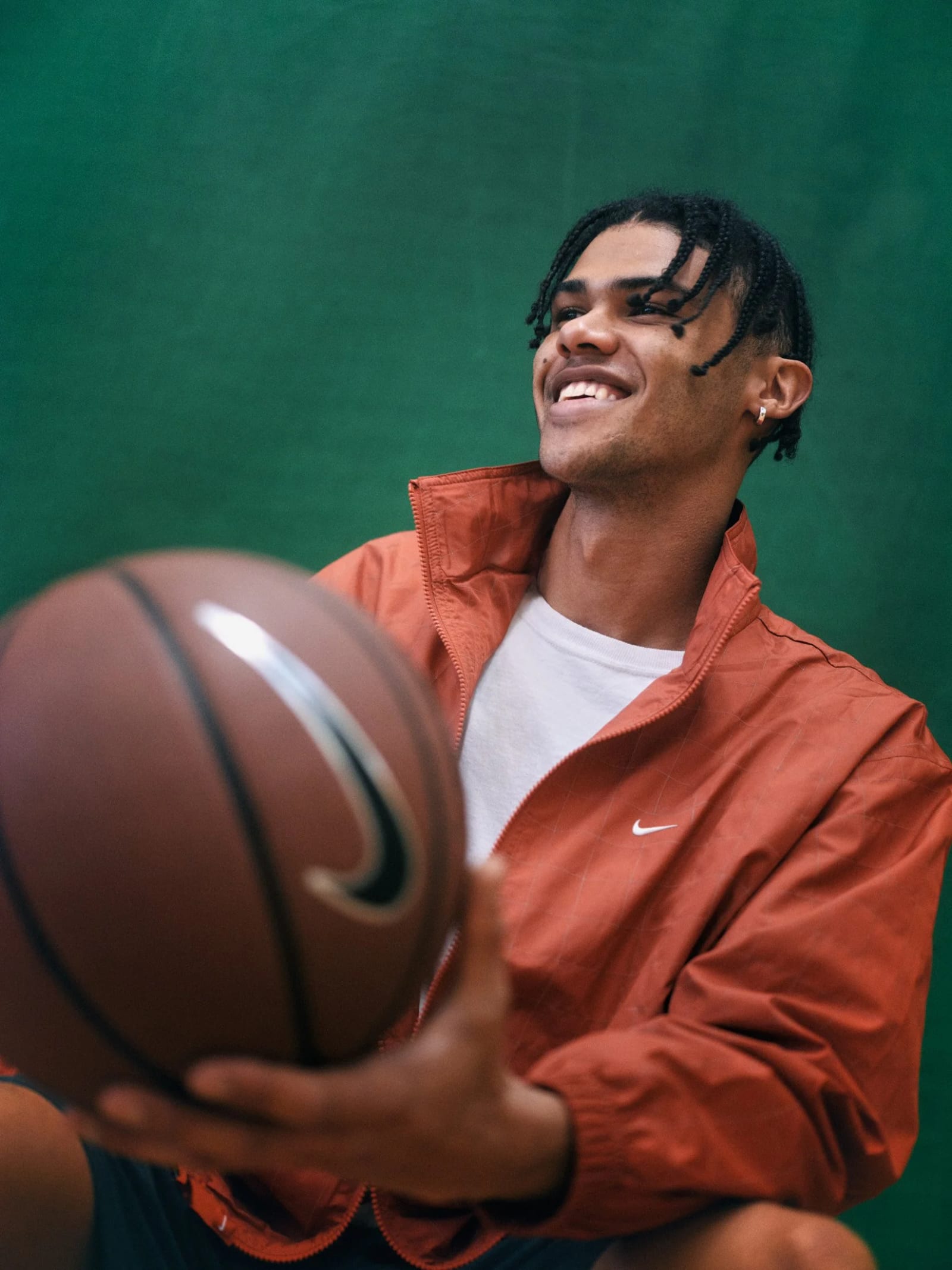 A smiling man with braids in a rust-orange Nike track jacket holding a basketball out toward the camera against a green wall.