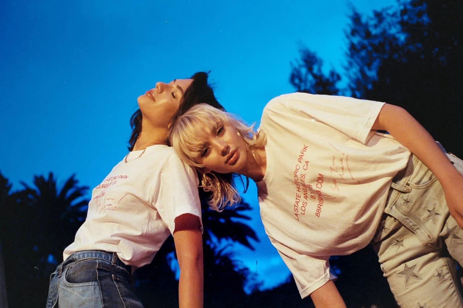 Two young women in printed white T-shirts leaning back to back under a deep blue evening sky and palm trees.