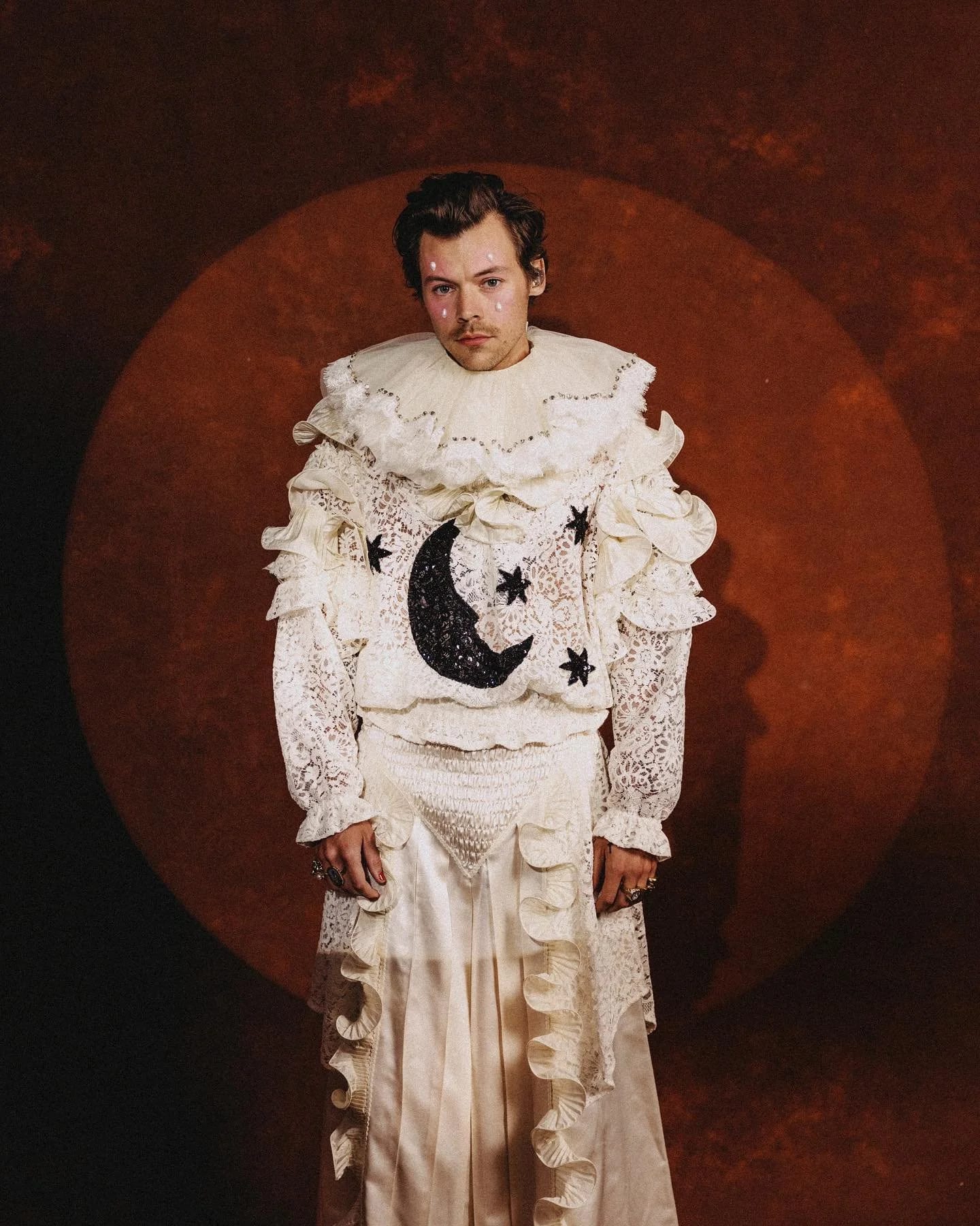 Harry Styles in a white ruffled costume with a moon and stars motif standing in a spotlit circle against a rust backdrop.