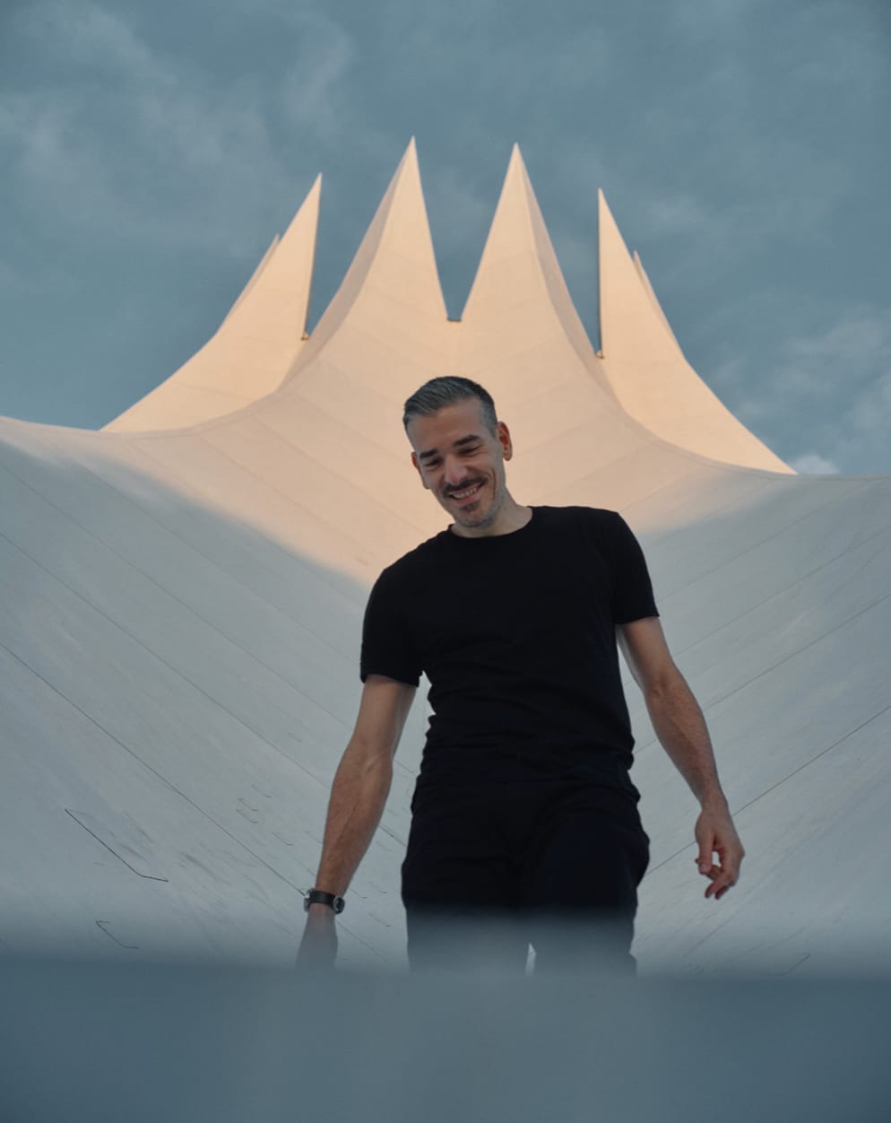 Anthony Kurtz in a black T-shirt standing in front of a large white tented roof with pointed peaks under a grey sky.