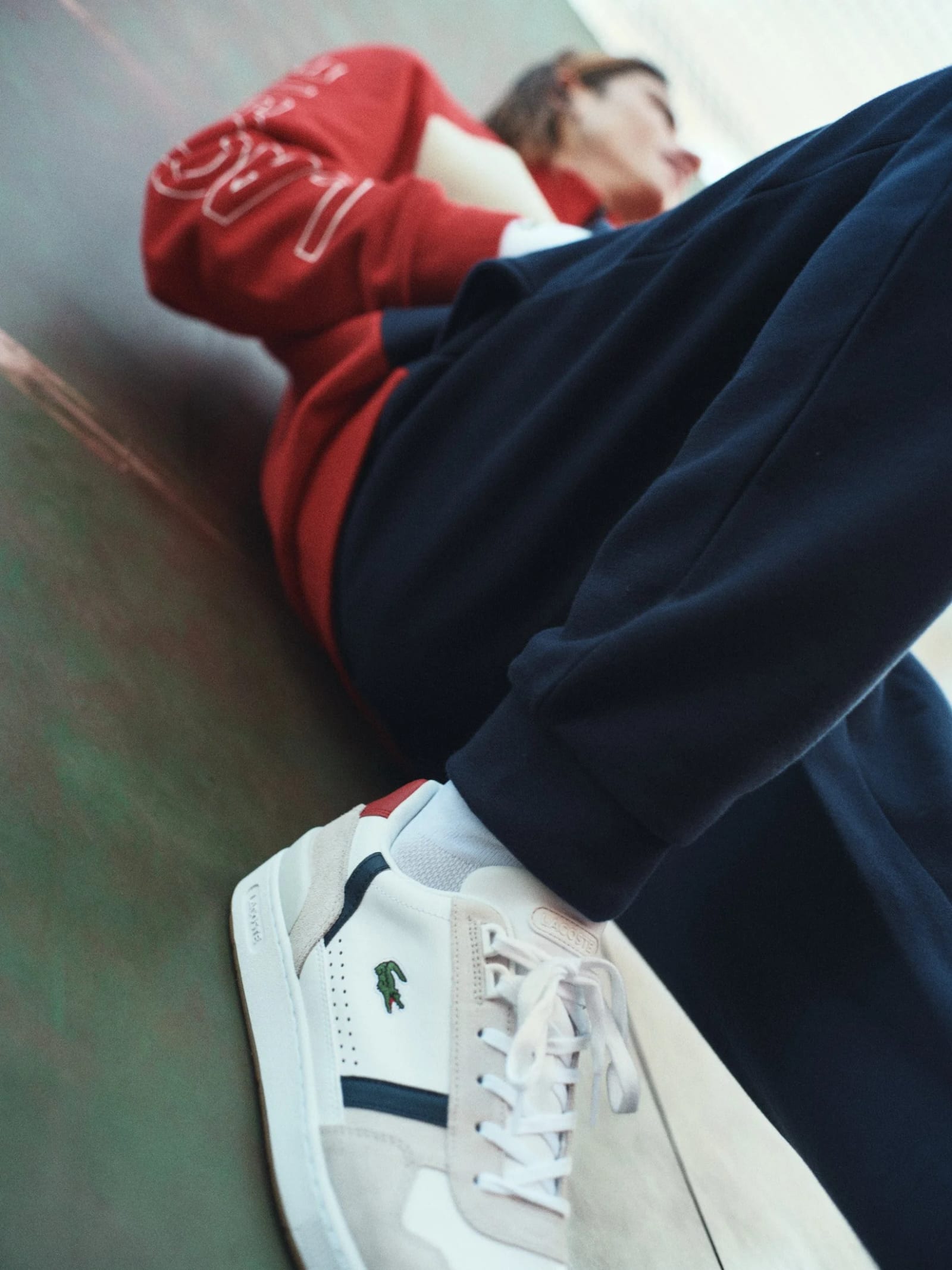 Low angle of white Lacoste trainers and navy trousers, a red-sleeved arm above.