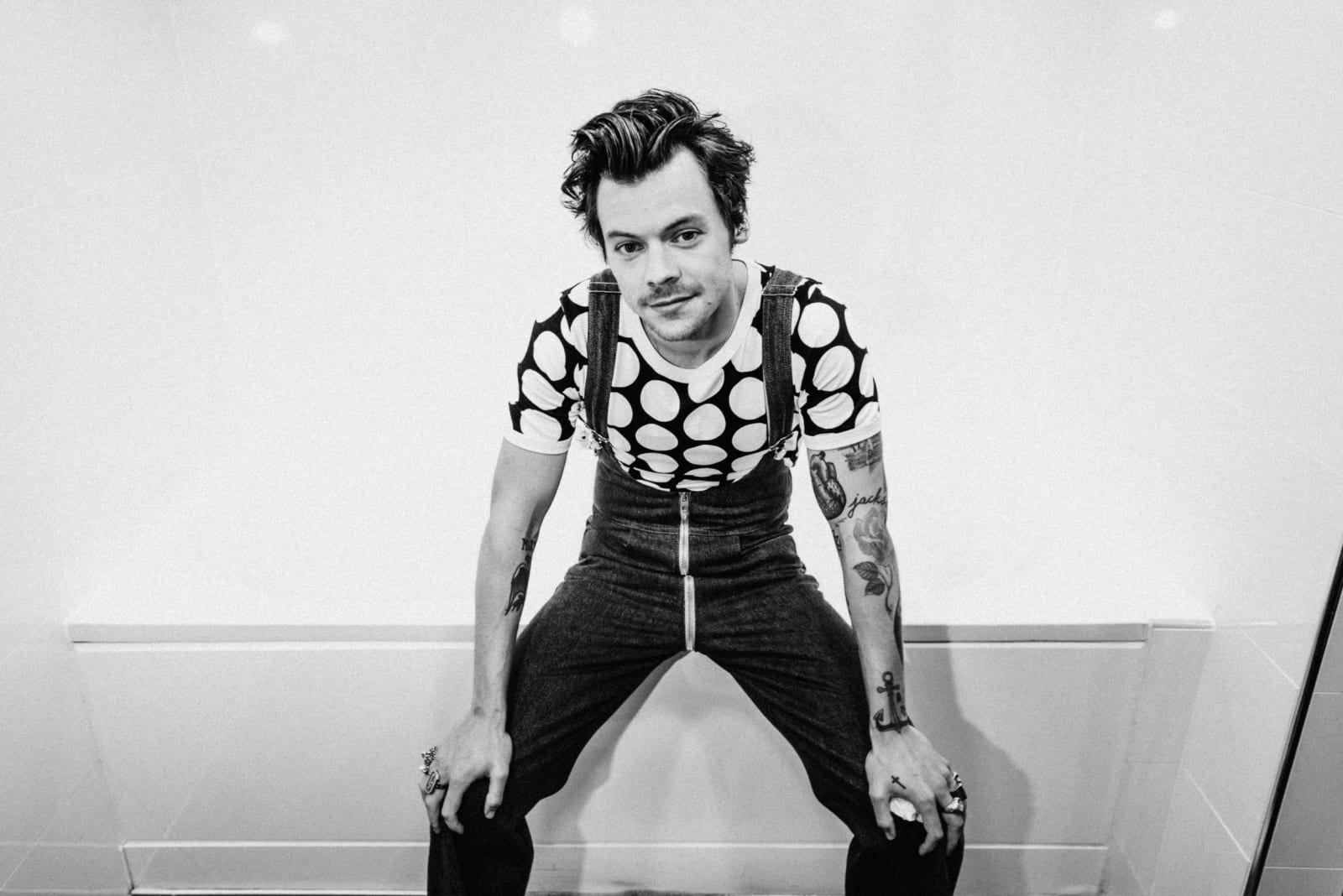 Black and white photograph of a man in a polka-dot top and dungarees crouching with his hands on his knees against a white wall.