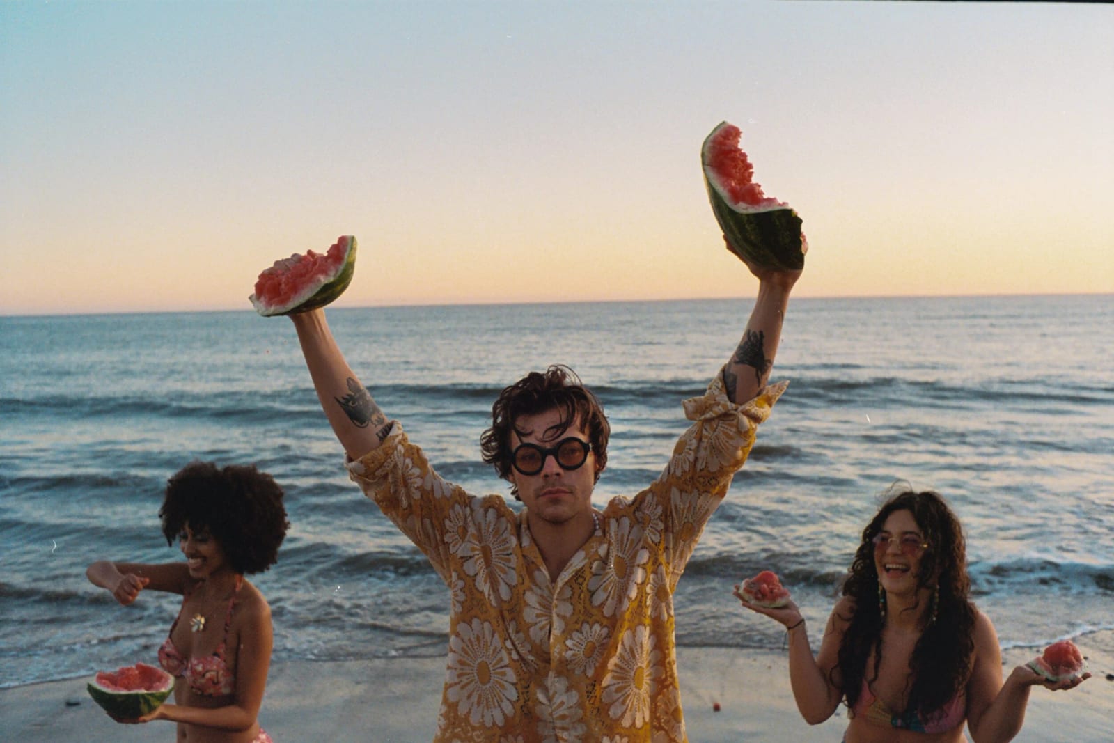 Harry Styles in a yellow floral shirt holding two slices of watermelon above his head at the shore, two women eating watermelon beside him.