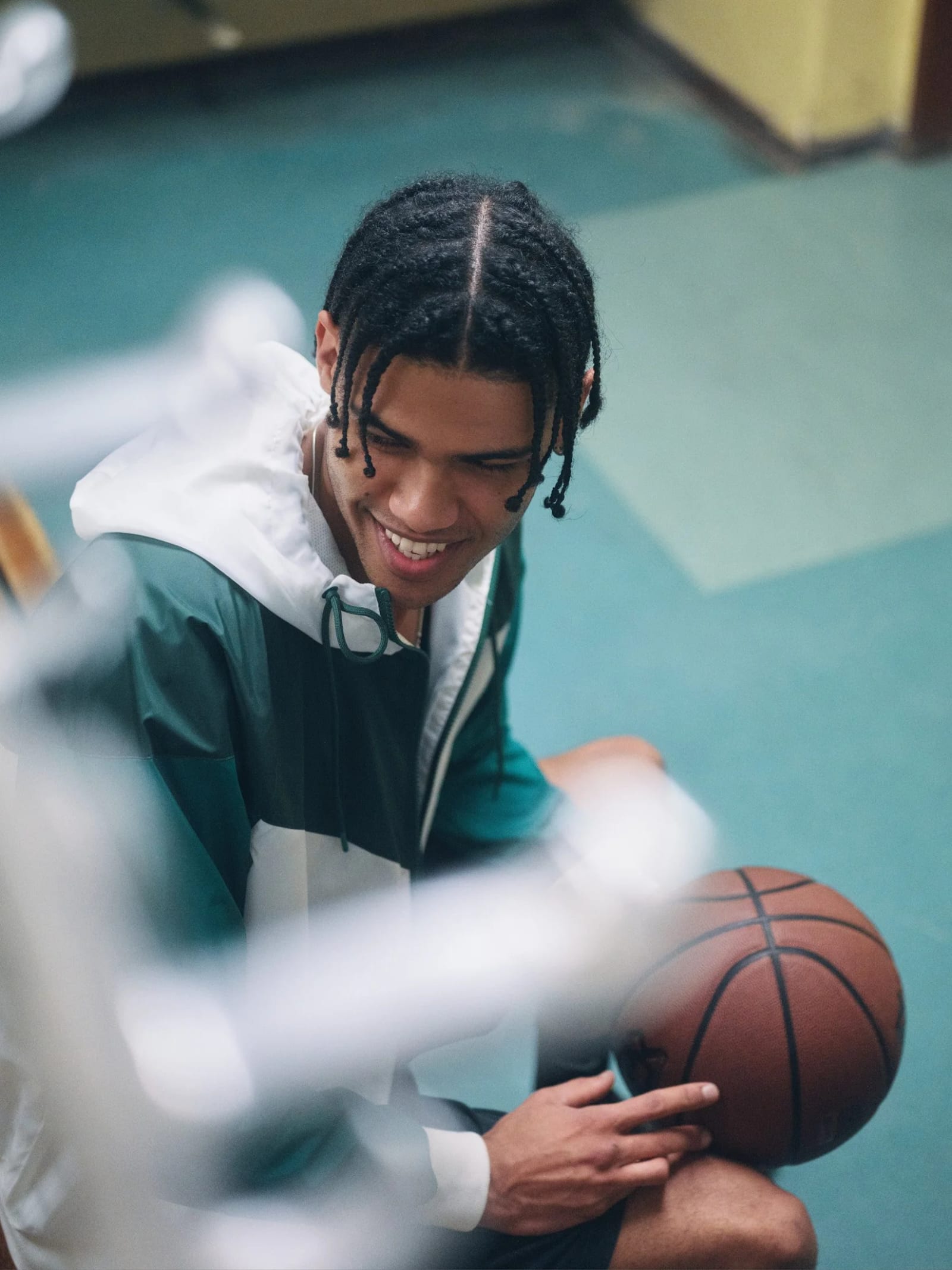 A smiling man with braids in a green and white track jacket crouching with a basketball, seen through blurred foreground.