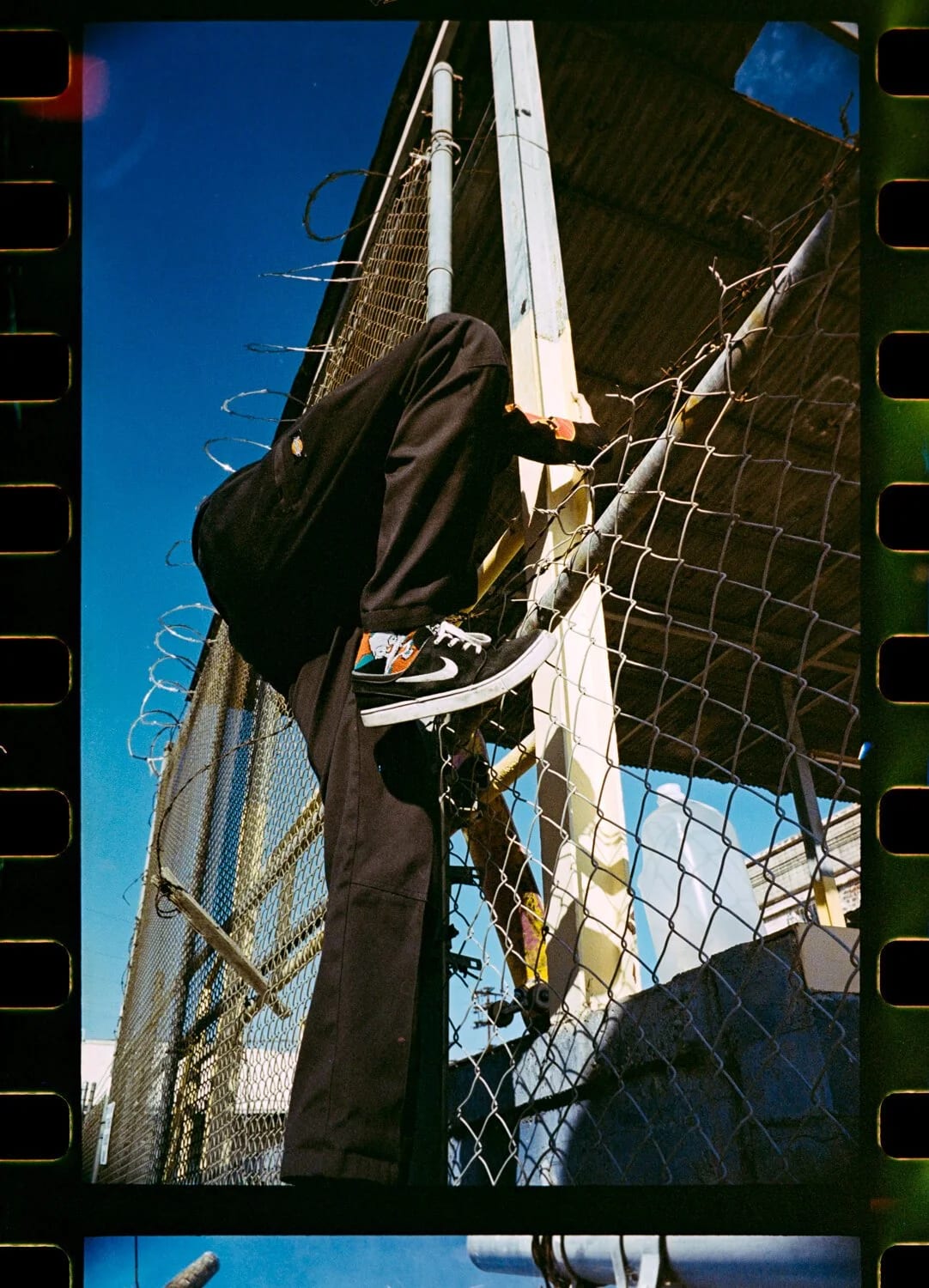 A film frame of a person in black climbing a chain-link fence topped with barbed wire against a blue sky.