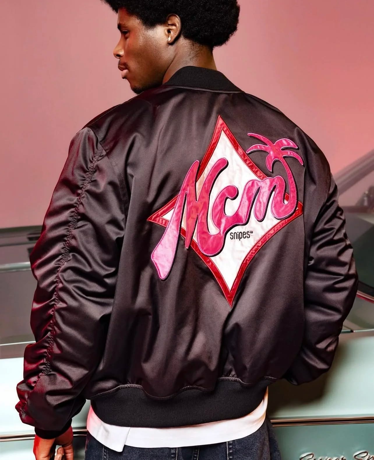 A man seen from behind in a black satin bomber jacket with a pink MCM diamond patch.