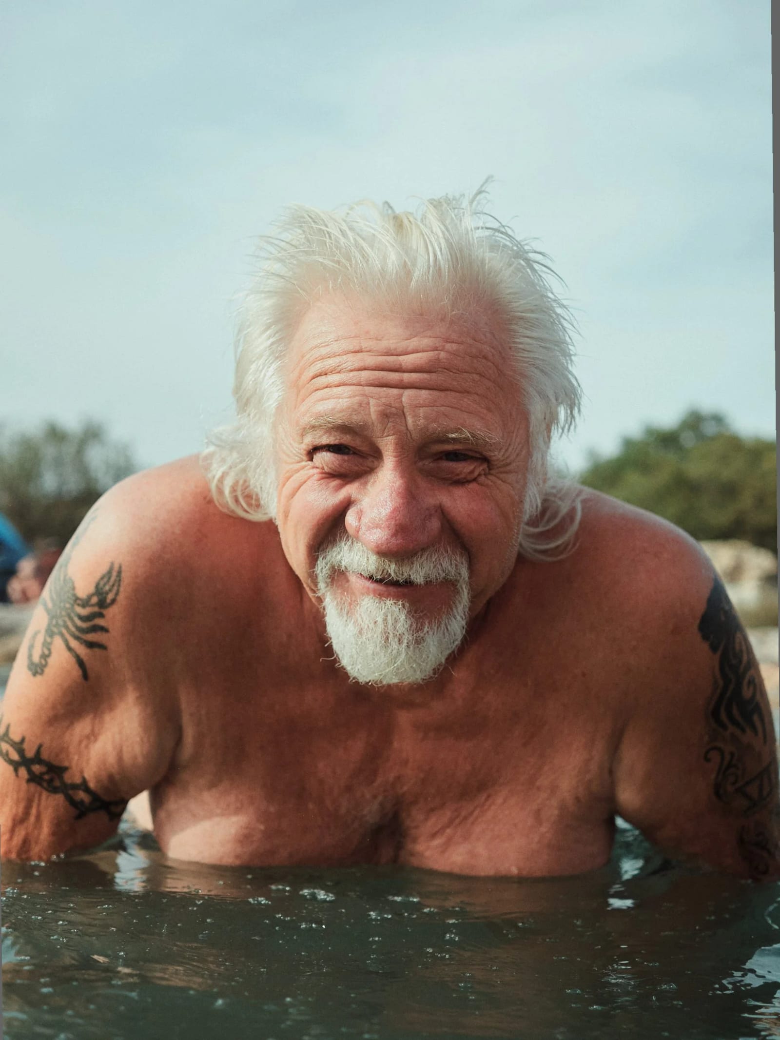 A white-haired man with a goatee and tattooed arms smiling from chest-deep water.