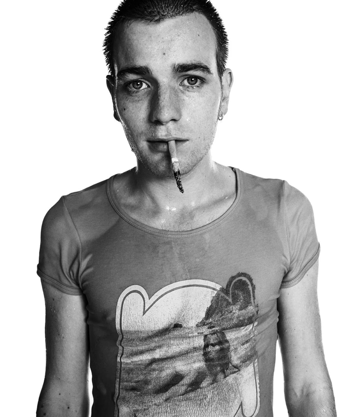 Black and white portrait of Ewan McGregor in a cropped printed T-shirt with a cigarette in his mouth, against white.