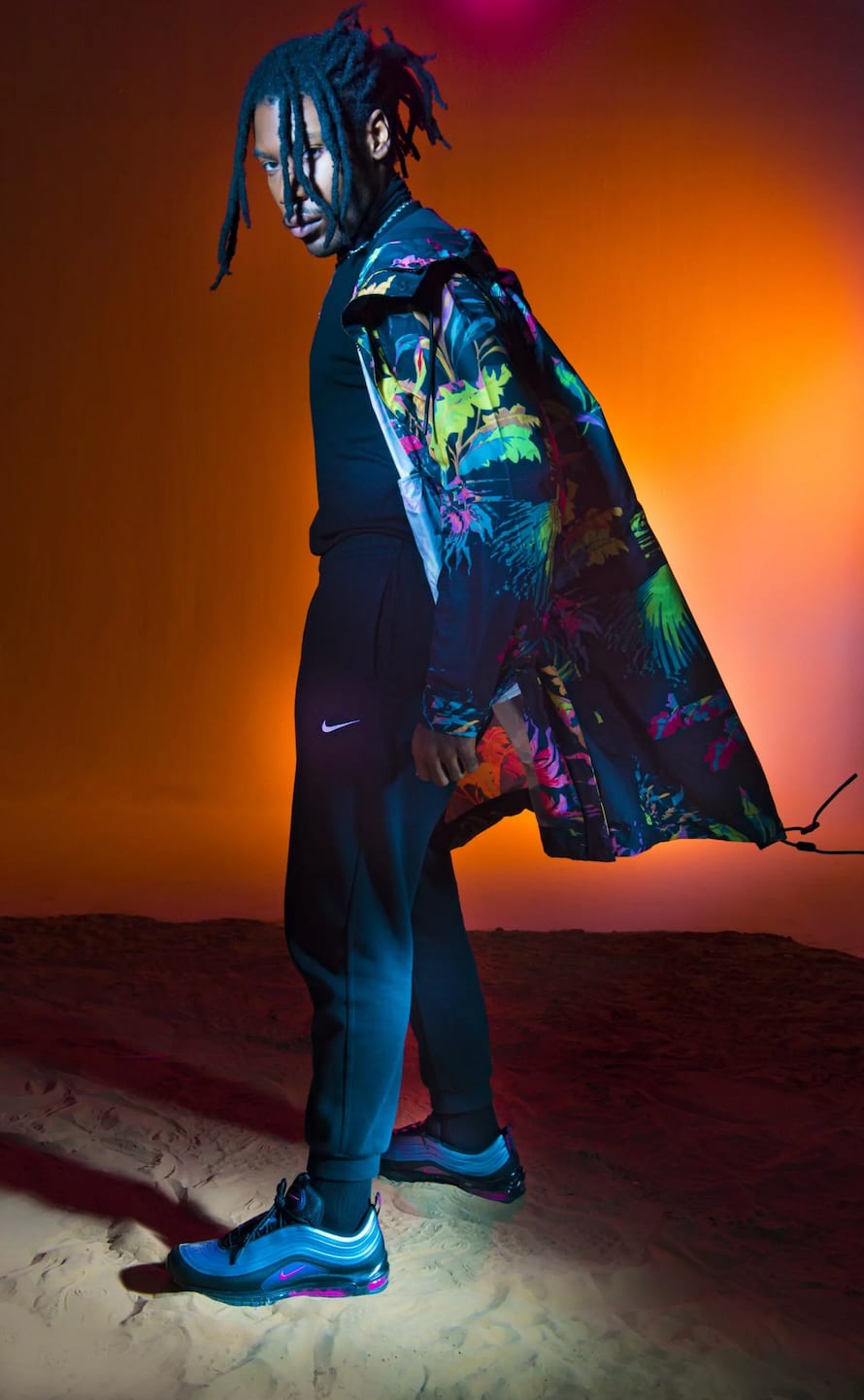 A man with locs in a floral-lined coat standing on sand, one arm holding the coat open, against orange.
