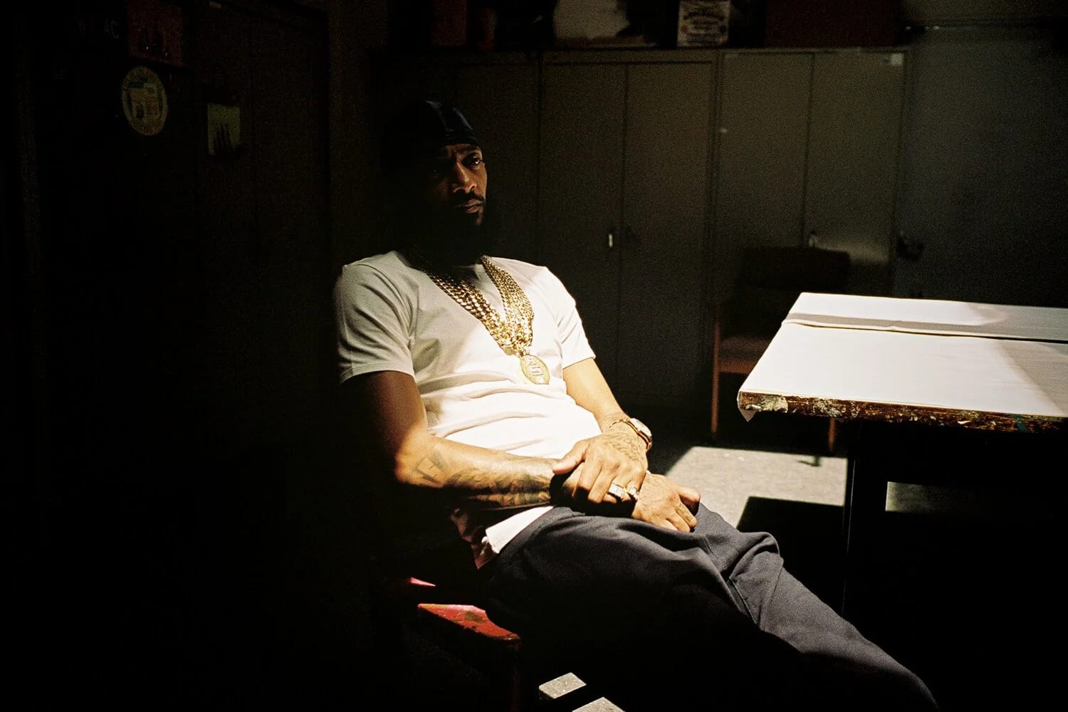 A man in a white T-shirt and gold chain sitting alone on a chair in a dim green room, a table beside him.