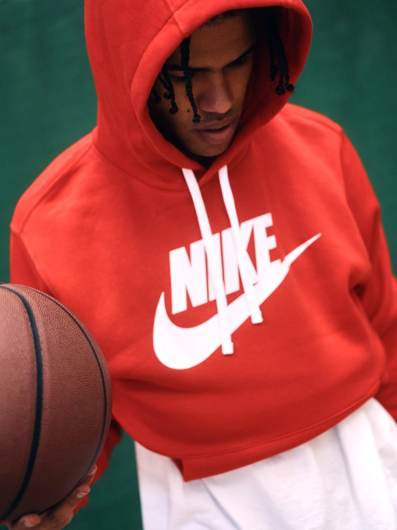 A man in a red Nike hoodie with the hood up looking down, a basketball in one hand, against a green wall.